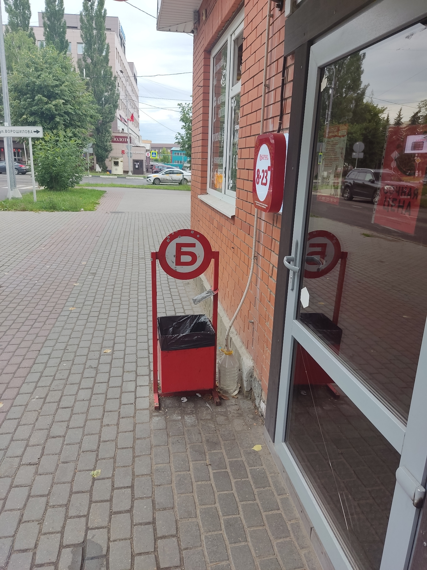 An outdoor scene in Serpukhov, Russia, features a sidewalk and the exterior of a brick building with large glass doors and windows. In the foreground, a red trash receptacle with a black liner stands on a paved sidewalk made of interlocking gray bricks. A red metal frame surrounds a circular sign with a white background and a large Cyrillic letter "Б" in red. To the right of the trash can, a plastic bottle filled with liquid is partially visible. The building's facade is constructed of red brick, and a round red sign with white lettering, indicating business hours (8-23), is mounted on the wall. Reflections in the glass doors show trees and the street. In the background, a multi-story building and more trees are visible. A street sign indicating "ул. ВОРОШИЛОВА" (Voroshilova Street) is also present. The sky is overcast.