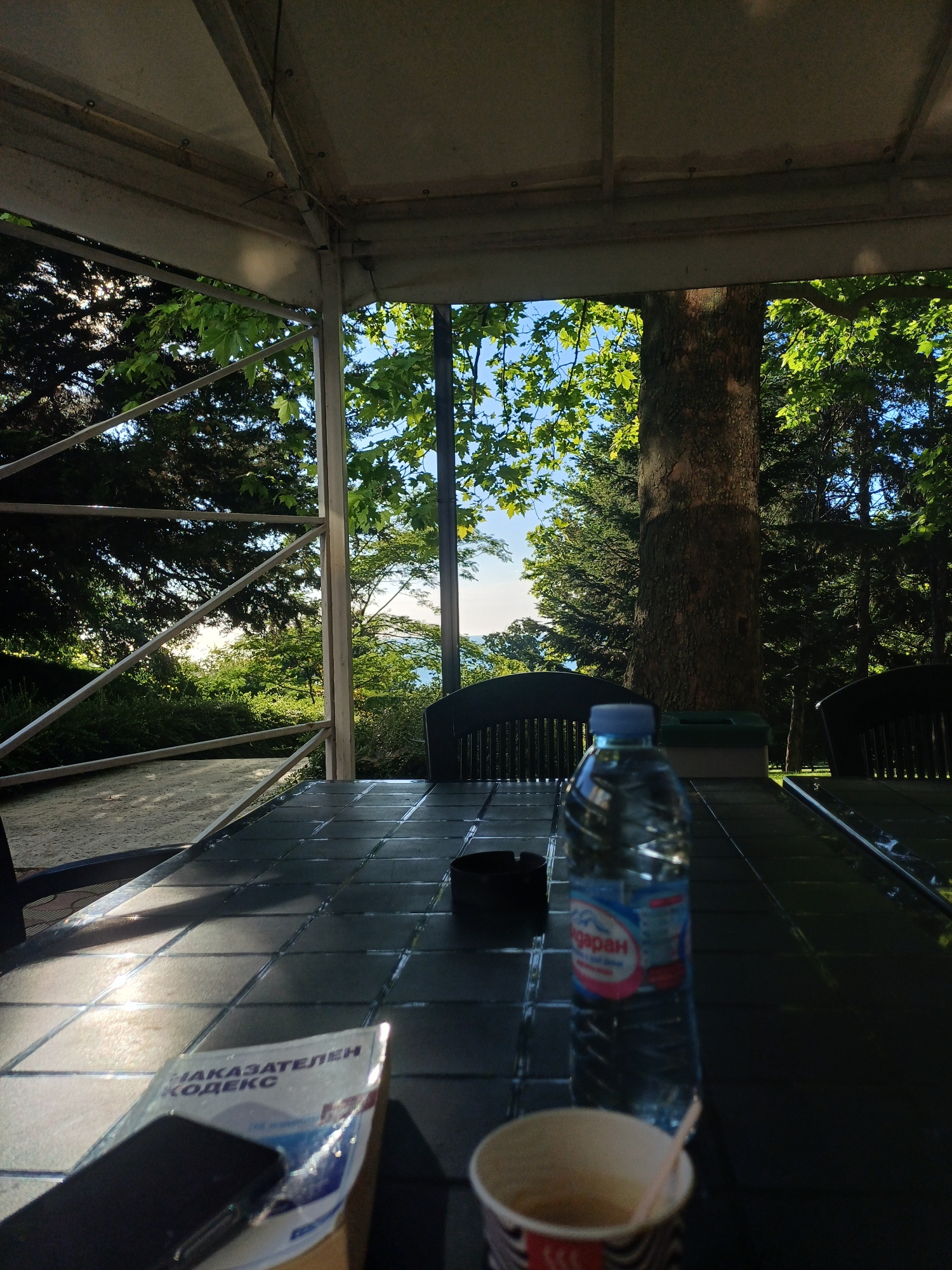 A table covered with a reflective, dark surface, possibly a patio table, is set with a plastic bottle of water, an ashtray, a phone, and a booklet with Cyrillic text that reads "НАКАЗАТЕЛЕН КОДЕКС." In the foreground, a disposable cup containing a beverage is also present. The table is situated outdoors under a white canopy, with sunlight filtering through lush green foliage and casting shadows on the table's surface. The background reveals a dense forest with a large tree trunk in the middle ground. The overall scene suggests a relaxed outdoor setting, potentially a park or a cafe in Varna, Bulgaria, during daylight hours.