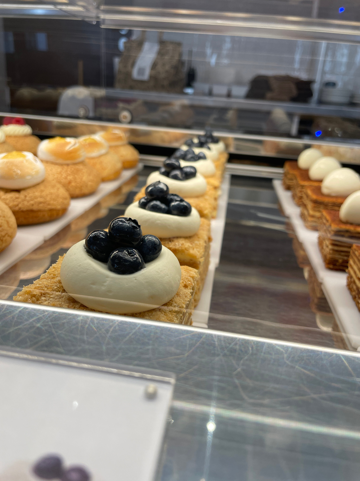 A close-up, eye-level shot shows a refrigerated display case filled with various pastries. In the foreground, a slice of mille-feuille topped with white cream and fresh blueberries is prominently displayed. To the left, several round pastries with white cream and blueberries are visible, some also adorned with a single blueberry or topped with what appears to be meringue. Further back, other pastries in different shapes and toppings are arranged in rows, including some with a glossy glaze and a bright red fruit topping. The display case has a clean, metallic finish and is illuminated, highlighting the sweet treats within. The background is blurred, but suggests other food items and shelving within the establishment, likely a bakery or patisserie in Belgrade, Serbia. The overall impression is one of a well-stocked and appealing dessert display.