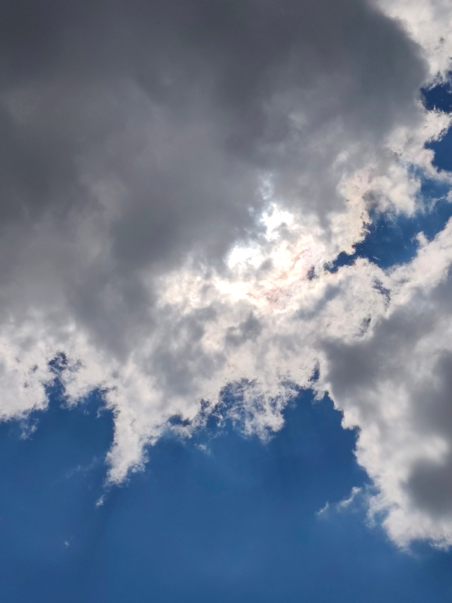 The image shows a cloudy sky with the sun partially visible through the clouds, creating a bright, diffused light. The clouds are a mix of dark gray and white, suggesting a dynamic weather system. The sky is a deep blue, indicating it's daytime. There are no people, objects, or specific city landmarks visible in this image, making it impossible to determine its location within Mykolaiv, Ukraine. The scene is a general atmospheric view.