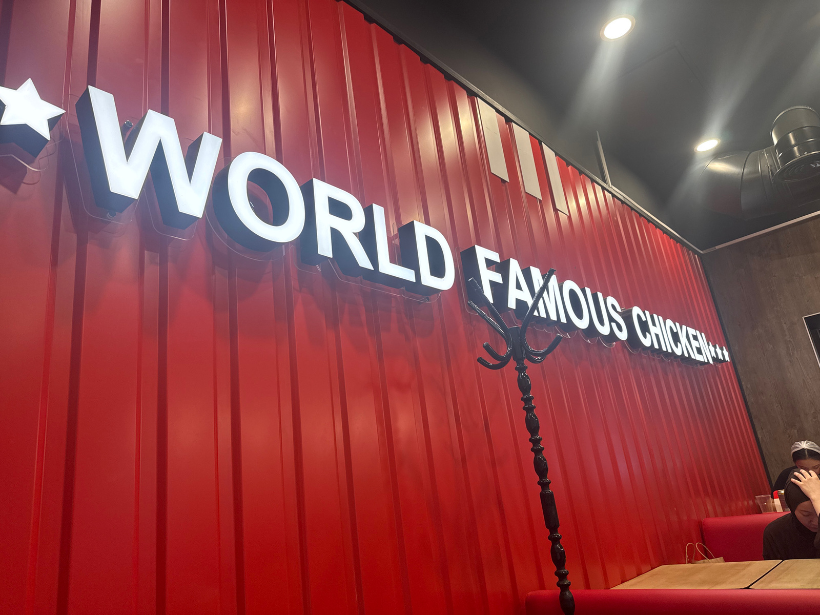 The image shows the interior of a restaurant in Almaty, Kazakhstan. The walls are a bright red corrugated metal, and a large illuminated sign reads "WORLD FAMOUS CHICKEN***". A black coat rack stands in front of the wall. In the foreground, a booth with red upholstery and a light brown tabletop is visible. Two people are seated in the booth, one wearing a dark hijab and the other with a white hair net. There are paper bags on the table. The ceiling is dark grey and features recessed lighting and a ventilation duct. The scene suggests a casual dining environment.