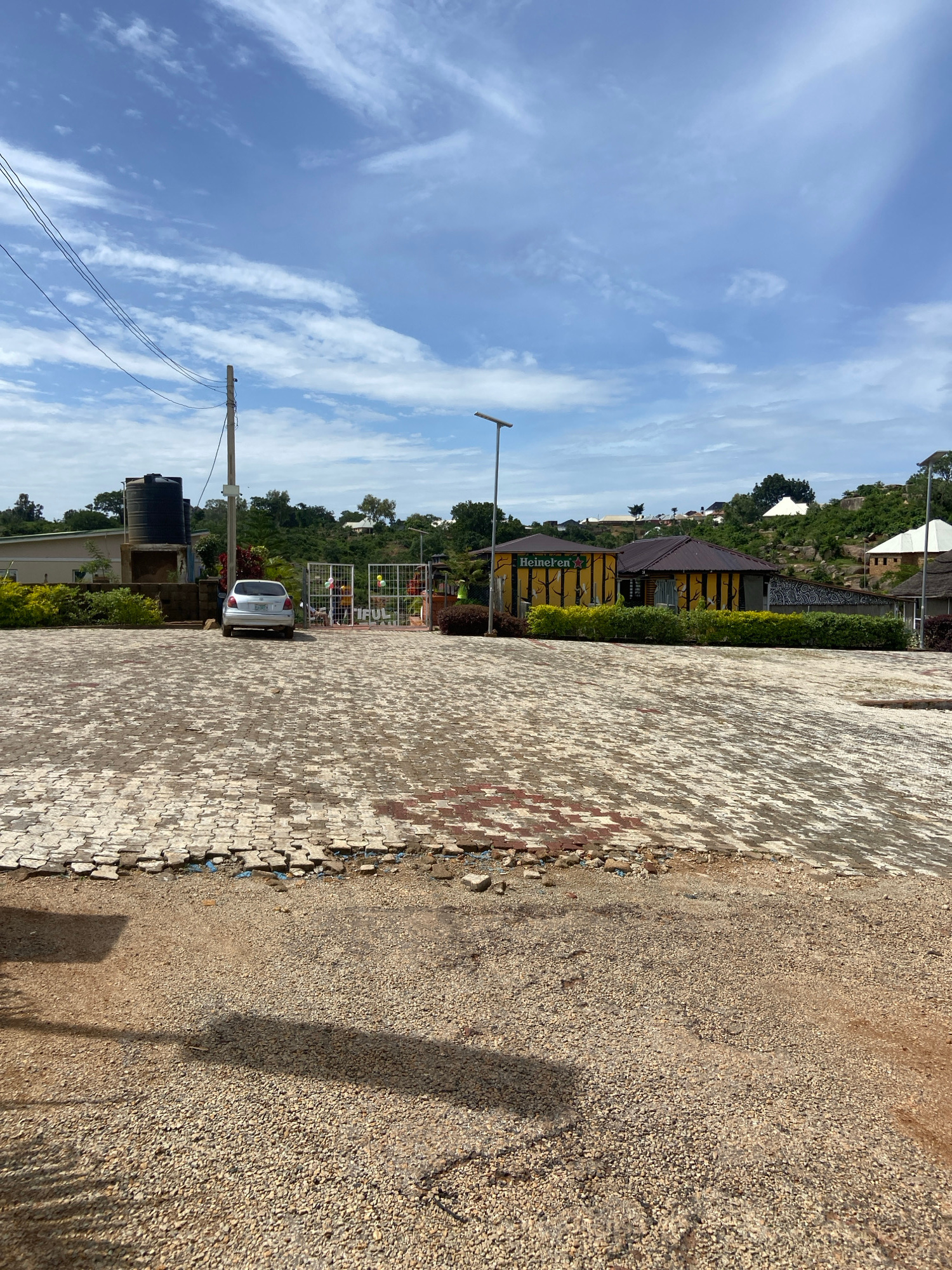 An exterior shot captures a paved area and a gravel lot in front of several buildings under a partly cloudy sky. The paved area, composed of interlocking bricks, shows signs of damage and disrepair in the foreground. A silver sedan is parked in the paved area, facing a gate with an inscription reading "TIFUL". Behind the car, a large black water tank is visible next to a building with green bushes. Further back, a yellow building with "Heineken" signage and tree motifs is situated on a hill, alongside other structures and vegetation. The environment suggests a location in Jos, Nigeria, possibly a commercial or hospitality establishment.
