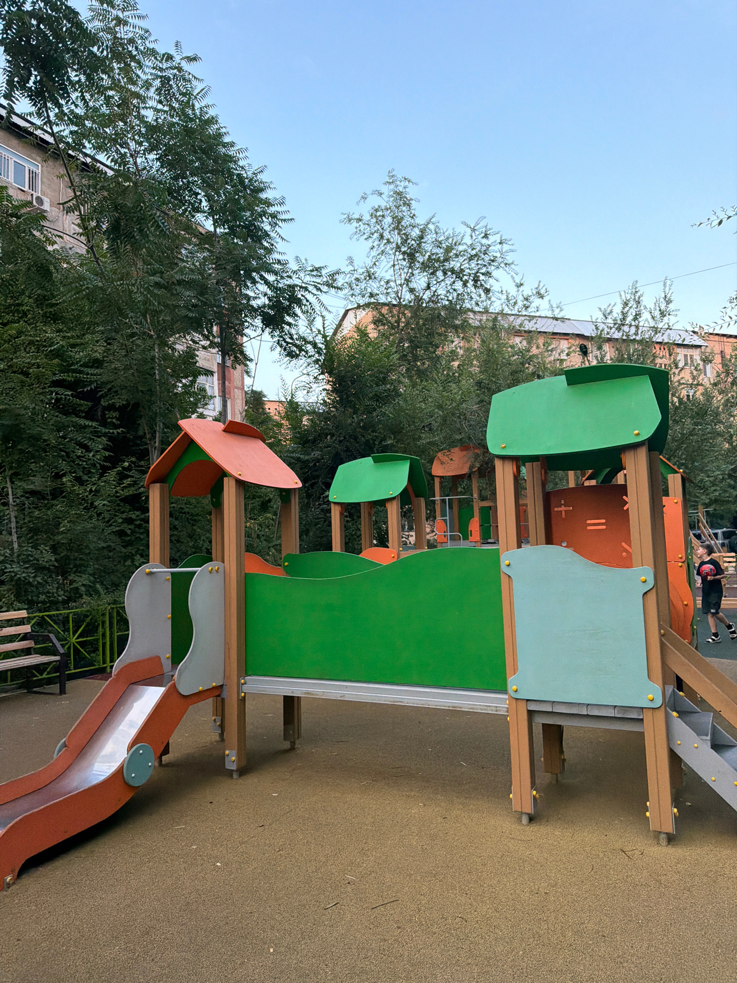 A children's playground is depicted, featuring a multi-level play structure with green and orange panels, a slide, and wooden supports. The ground is a textured, tan-colored surface. In the background, residential buildings and lush trees are visible, suggesting an urban setting. A young boy, wearing shorts and a t-shirt, is walking to the right, holding a red ball. The sky is clear and blue. The scene appears to be in a residential area of Shymkent, Kazakhstan.