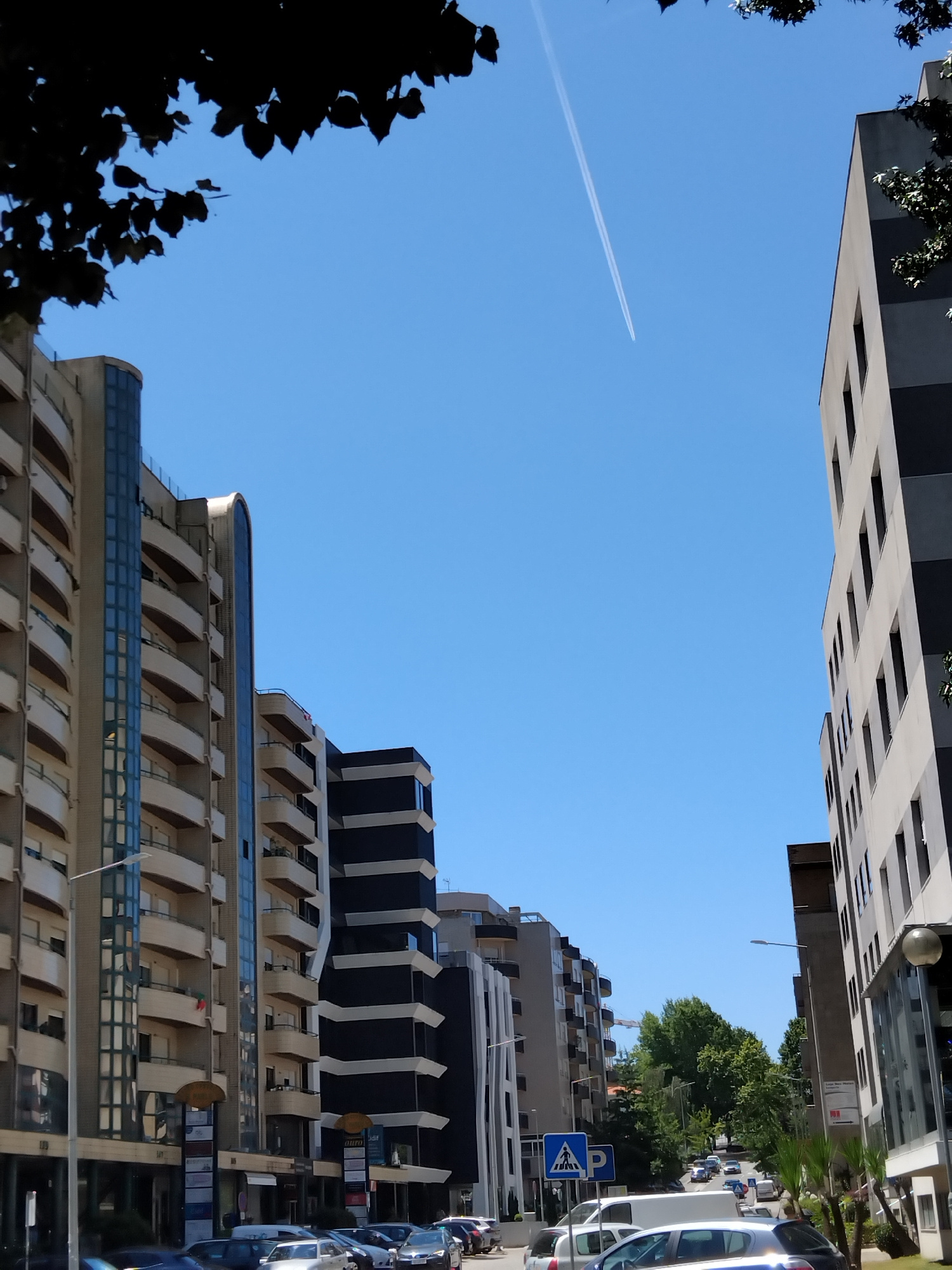 The image captures a street scene in Vila Nova de Famalicão, Portugal. The perspective is from street level, looking upwards towards a clear blue sky with a contrail from an airplane. On the left, a modern apartment building with balconies and glass elements dominates the frame. To the right, another contemporary building with a dark and light gray facade is visible. In the background, a row of buildings extends down the street, interspersed with trees. The street itself is lined with parked cars and some palm trees. A pedestrian crossing sign and a parking sign are present on the right side of the street. The overall impression is one of an urban environment on a bright, sunny day.