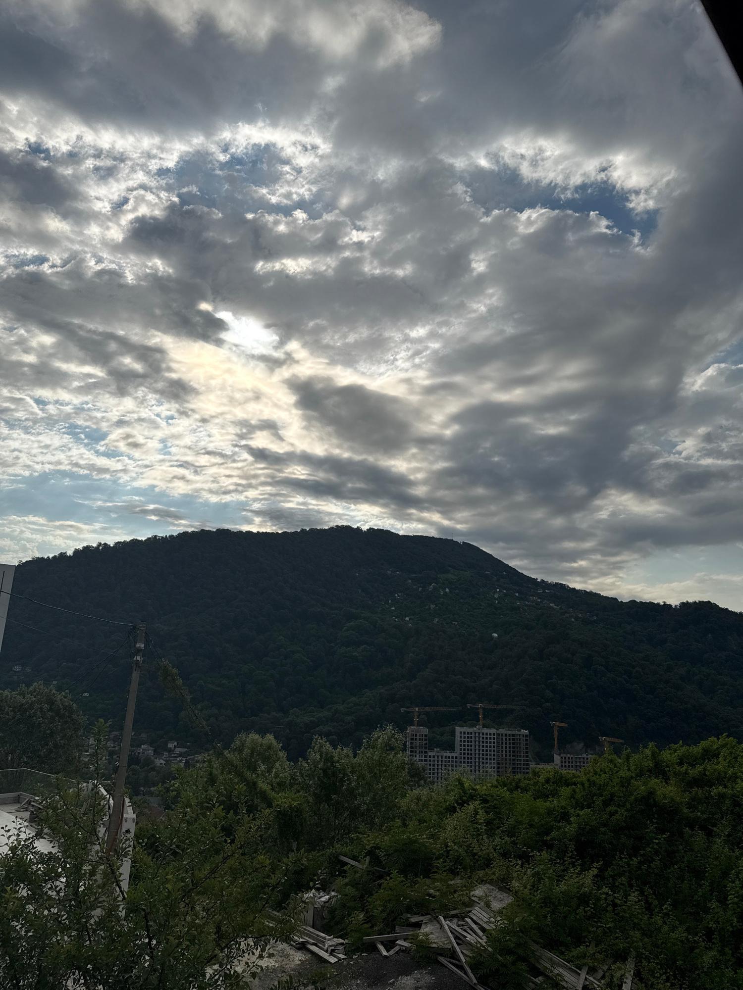 A vertical, slightly low-angle shot captures a cloudy sky above a densely forested mountain, with construction cranes and buildings visible in the middle ground. The sky is filled with dramatic, grey clouds, with patches of blue sky and sunlight breaking through. The mountain, covered in dark green trees, dominates the lower half of the frame. In the middle ground, a complex of multi-story buildings, likely under construction, is visible, with several yellow construction cranes towering over them. The foreground features lush green foliage and what appears to be construction debris, including wooden planks. The image appears to be taken in the vicinity of Baranovka, Russia, given the context provided. There are no people or animals visible in the scene.