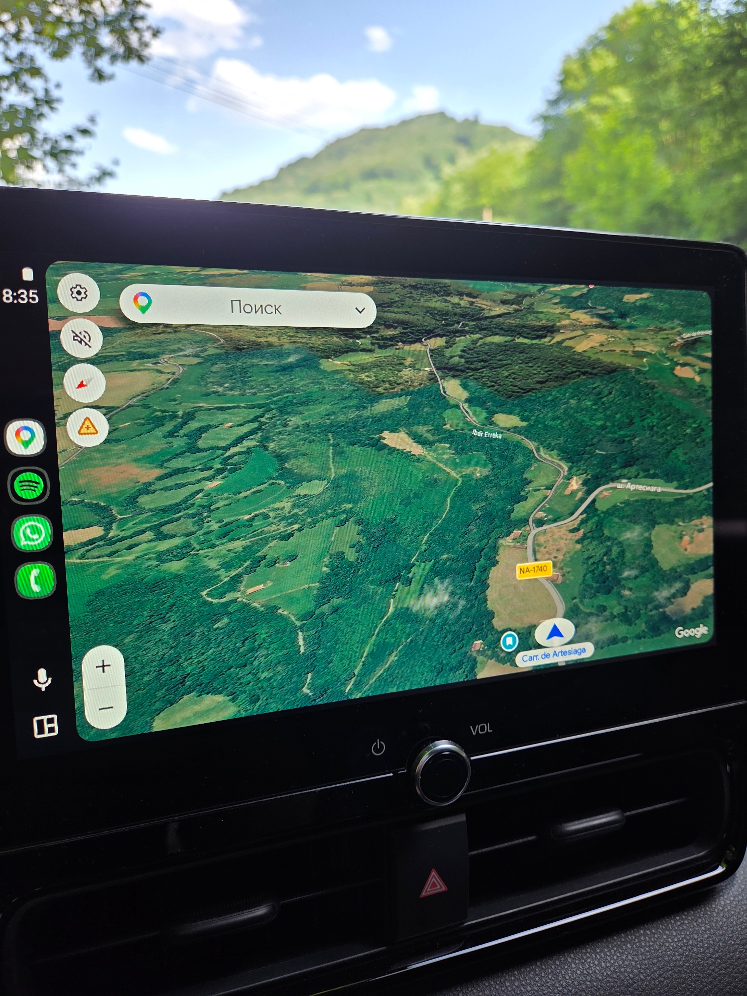 The image shows a car's dashboard display showing a Google Maps navigation screen. The map displays a satellite view of a rural, mountainous area with green forests, fields, and a winding road. The road has a yellow sign labeled "NA-1740". Text on the map indicates the presence of roads labeled "Ibâr Erreka" and "ш. Артесиага", and a destination labeled "Carr. de Artesiaga". The display also shows icons for Google, Spotify, WhatsApp, and a phone. A time of "8:35" is visible in the top left corner. The display is surrounded by the car's interior, including air vents and buttons. The background outside the car shows a bright blue sky with clouds and a green mountain range. The scene is likely captured while the car is parked or driving in the region of Baztan, Spain, given the road signage and geographical features depicted in the map.