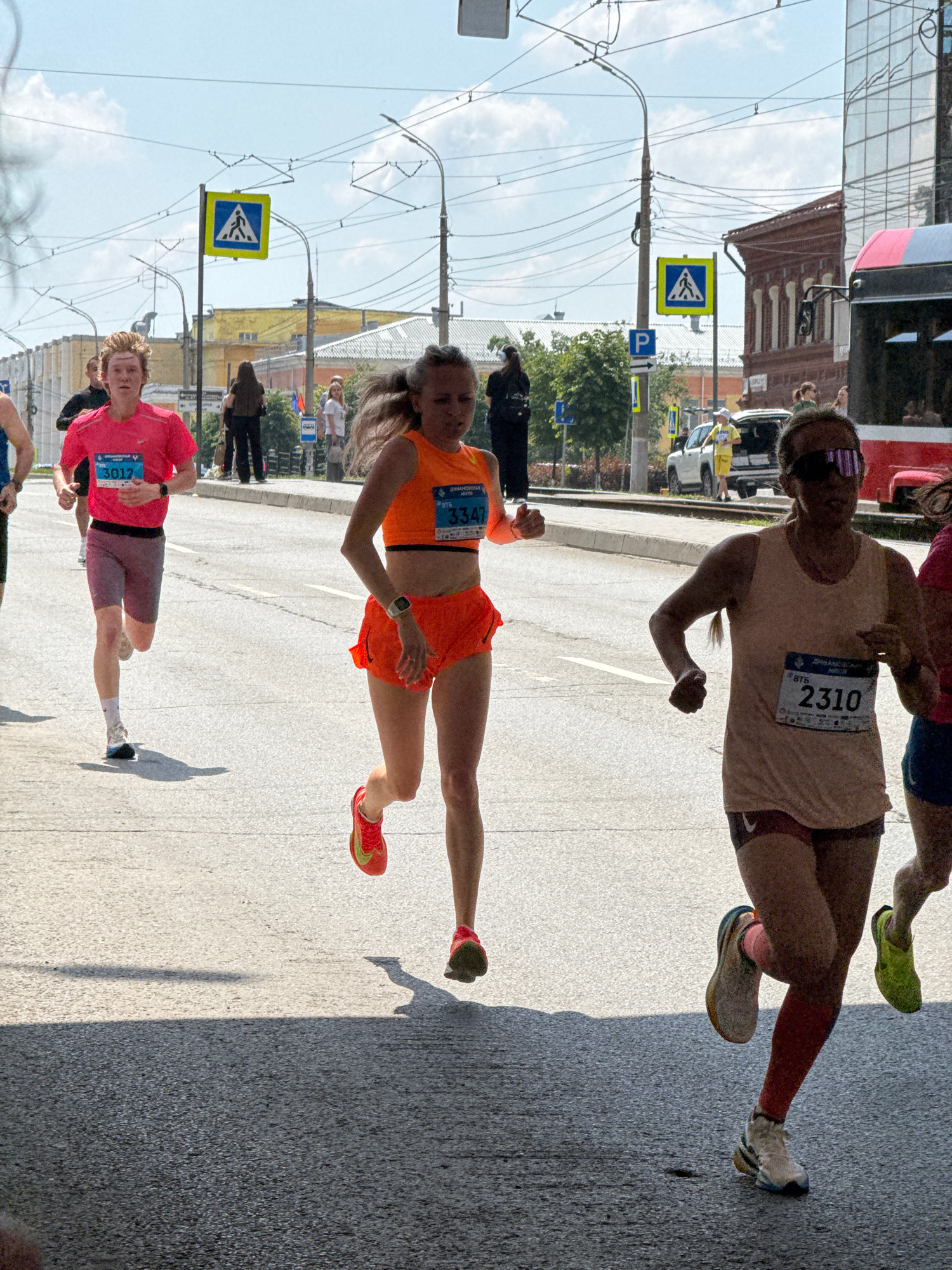 The image depicts a road race taking place on a sunny day in Izhevsk, Russia, likely on Pushkinskaya Street, given the architectural style of the buildings and the presence of tram lines. Four female runners are prominently featured in the foreground, each wearing athletic attire and bib numbers. The runner on the far left wears a pink t-shirt and grey shorts, with bib number 3012. The central runner is in an orange crop top and orange shorts, displaying bib number 3347. To the right of her is a runner in a light brown tank top and dark shorts, with bib number 2310. Another runner, partially visible on the far right, is in a pink top and dark shorts. The background shows streetlights, overhead wires, buildings with red brick and modern glass facades, parked vehicles, and pedestrians observing the race. Pedestrian crossing signs are visible, indicating the race route passes through an urban area. The road surface is asphalt.