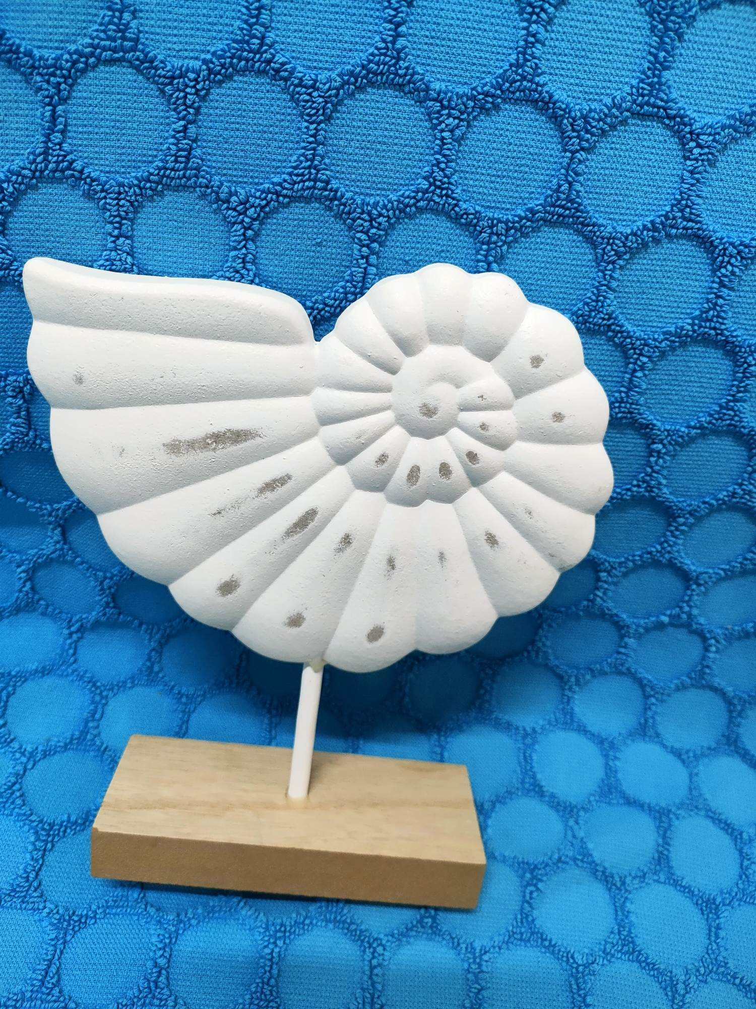 A white, seashell-shaped decorative object is displayed on a light-colored wooden base against a textured blue background. The seashell has a spiral design with indentations and some gray marks. The blue background appears to be a fabric with a honeycomb-like pattern. The object is positioned slightly to the left of the center of the frame. There are no people or animals visible. The scene does not provide any information related to the city of Nosivka, Ukraine.