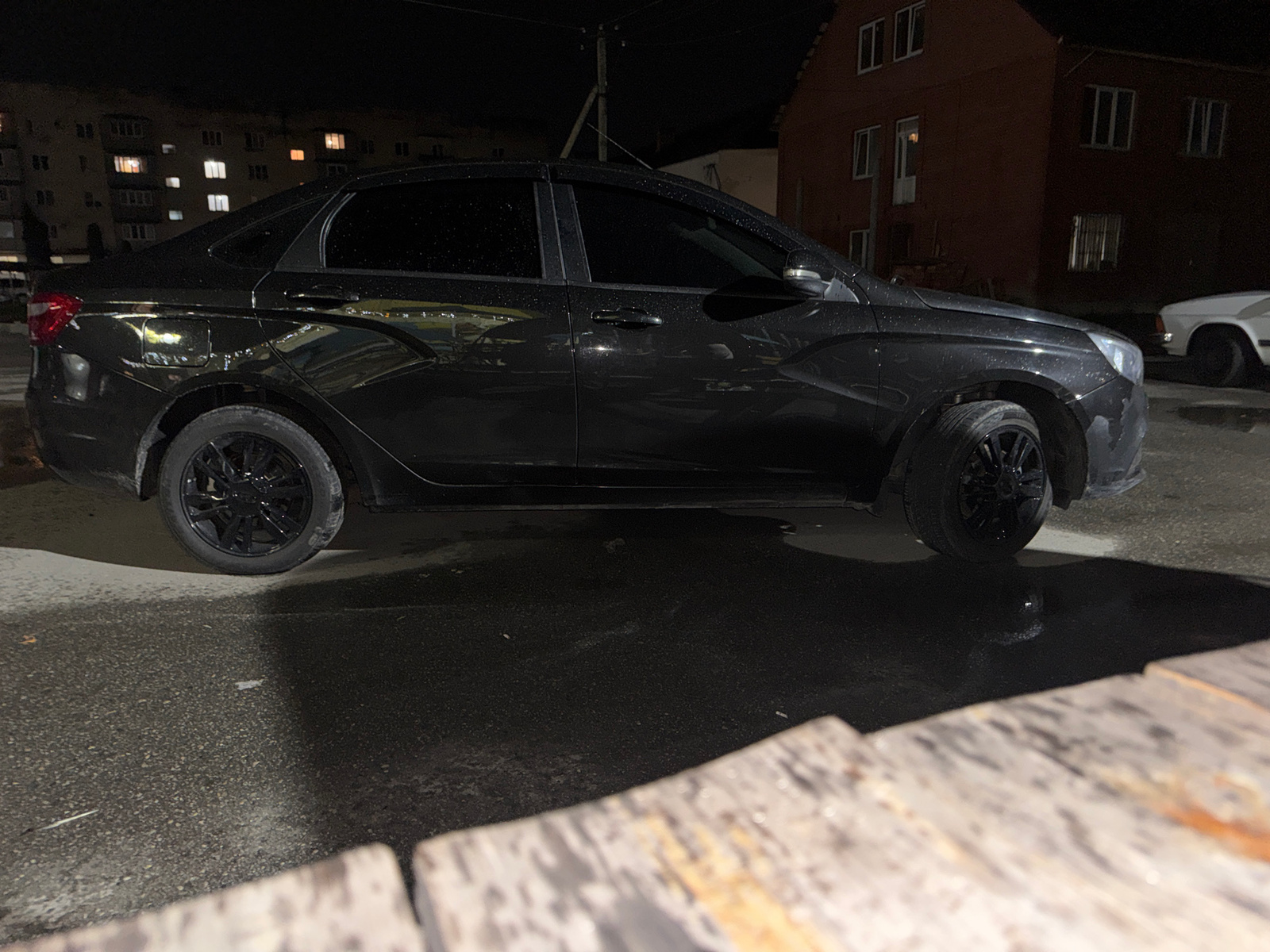 An eye-level, medium shot shows a black sedan parked on a wet asphalt surface at night. The car is positioned centrally in the frame, with its front facing to the right. Reflections of lights from a nearby building are visible on the car's surface, indicating it has recently rained. The background features a multi-story brick building with several lit windows. To the right of the sedan, a white vehicle is partially visible, also parked. The foreground is occupied by a blurred wooden structure, likely a fence or railing, out of focus. The lighting suggests artificial light sources from the buildings and possibly streetlights.