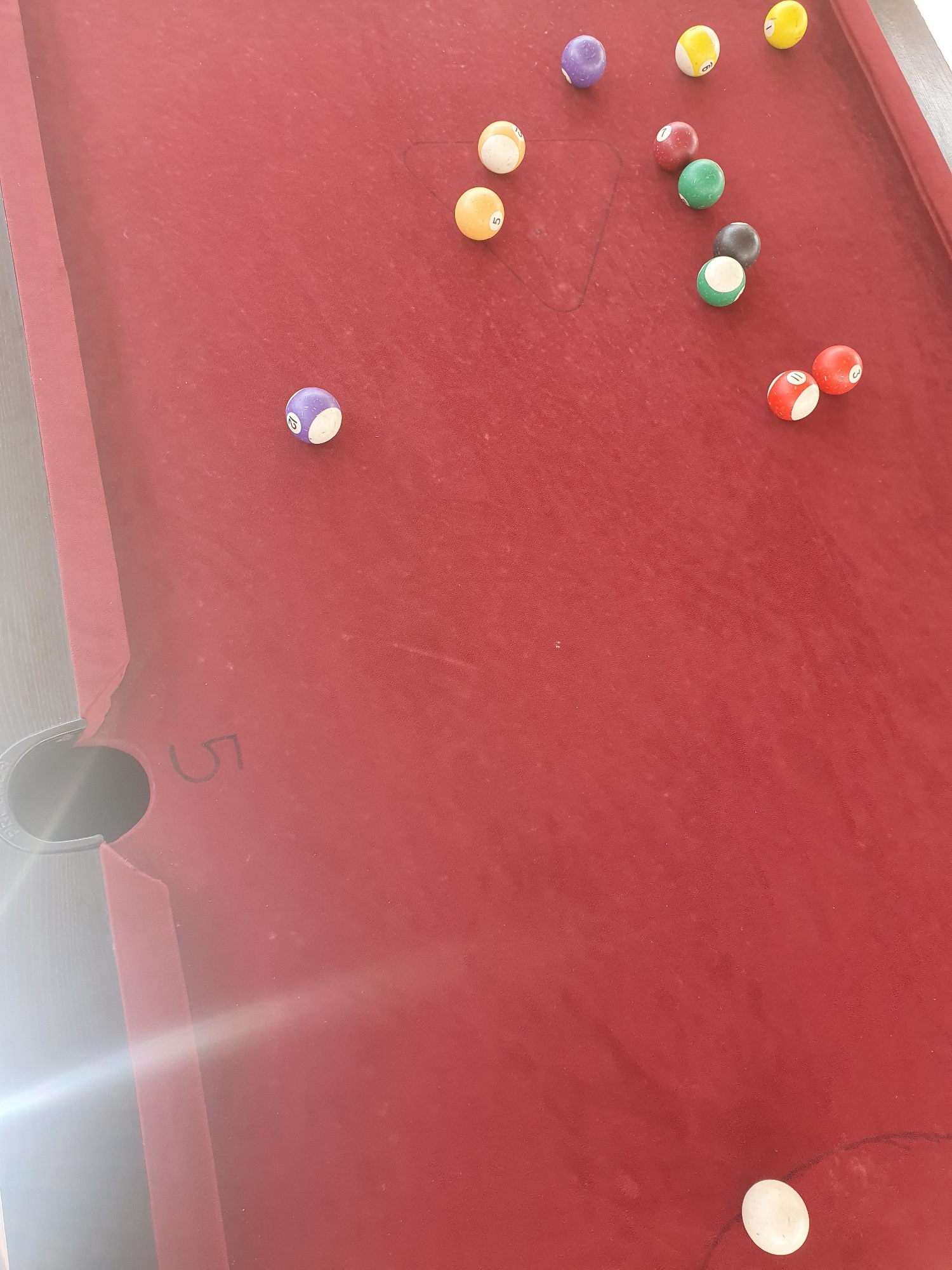 A pool table with several billiard balls is shown. The table is covered in red felt. A white cue ball is at the bottom right of the image. There are several numbered balls scattered across the table, including a purple ball with the number 12, an orange ball with the number 5, a yellow ball with the number 9, a red ball with the number 7, a green ball, a black ball, a green and white ball, a red ball with the number 11, and a red ball with the number 3. The pool table is positioned outdoors, possibly in a courtyard or patio, with a bright light source casting lens flare on the left side of the image. The number "5" is visible on the felt of the table near the bottom left pocket. The environment is not specifically identifiable as Lokoja, Nigeria, but appears to be an outdoor recreational area. There are no people or actions depicted in the image.