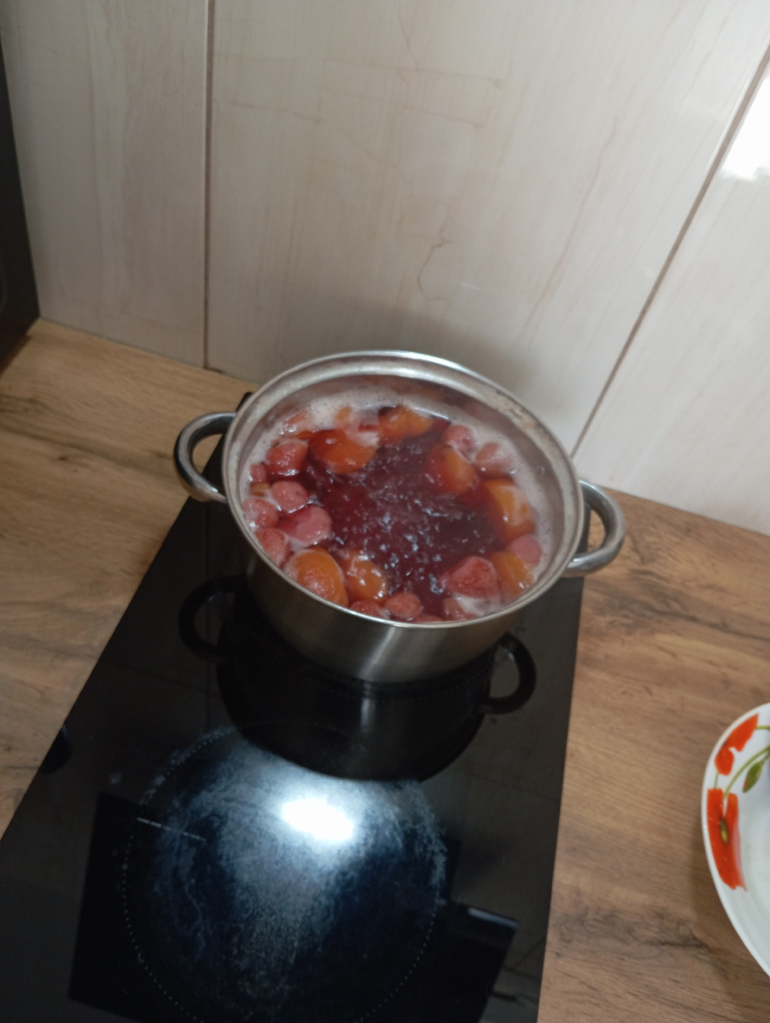 A metal pot filled with boiling fruit, likely strawberries and possibly plums or cherries, sits on a black glass stovetop. The fruit is submerged in a dark red liquid that is bubbling and frothing. The stovetop has circular heating elements indicated by dotted lines. The kitchen counter is made of wood-look laminate, and the backsplash is tiled with a light-colored, slightly textured material. A plate with a floral pattern, featuring red poppies, is partially visible on the right side of the counter. The image appears to have been taken in a residential kitchen.