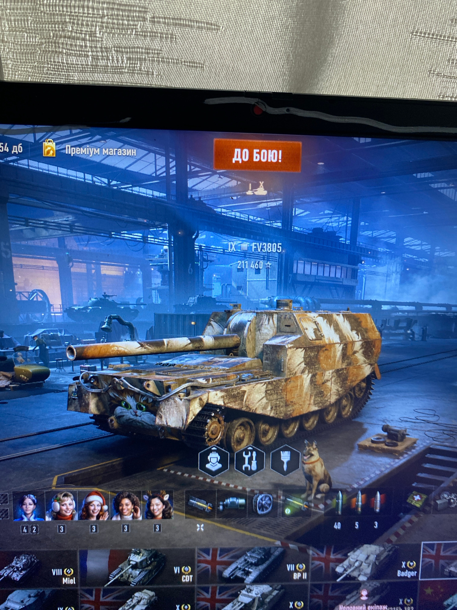 The image depicts a screenshot from a video game, likely a tank combat simulation given the prominent display of a tank and related icons. The scene shows a virtual garage or hangar environment with industrial elements like large metal structures and machinery.
In the foreground, a camouflaged tank with a distinctive cat decal on its front armor is the primary subject. To the left of the tank, there is an interface displaying what appear to be crew portraits, possibly representing different crew members with assigned numbers. Below this, a row of selectable tanks is visible, each identified by a tier, country flag (including French and British), and name.
In the upper portion of the screen, there are game-related buttons. "Преміум магазин" (Premium Shop) and "ДО БОЮ!" (To Battle!) are clearly visible, suggesting typical game progression options. The text "IX FV3805" indicates the specific tank model displayed is tier IX, FV3805.
The background shows a dimly lit, industrial interior with multiple other tanks and structures, suggesting a virtual game environment. There is no indication of a real-world location such as Szczecin, Poland, as this is a digital rendering. The image is a still capture from a screen.