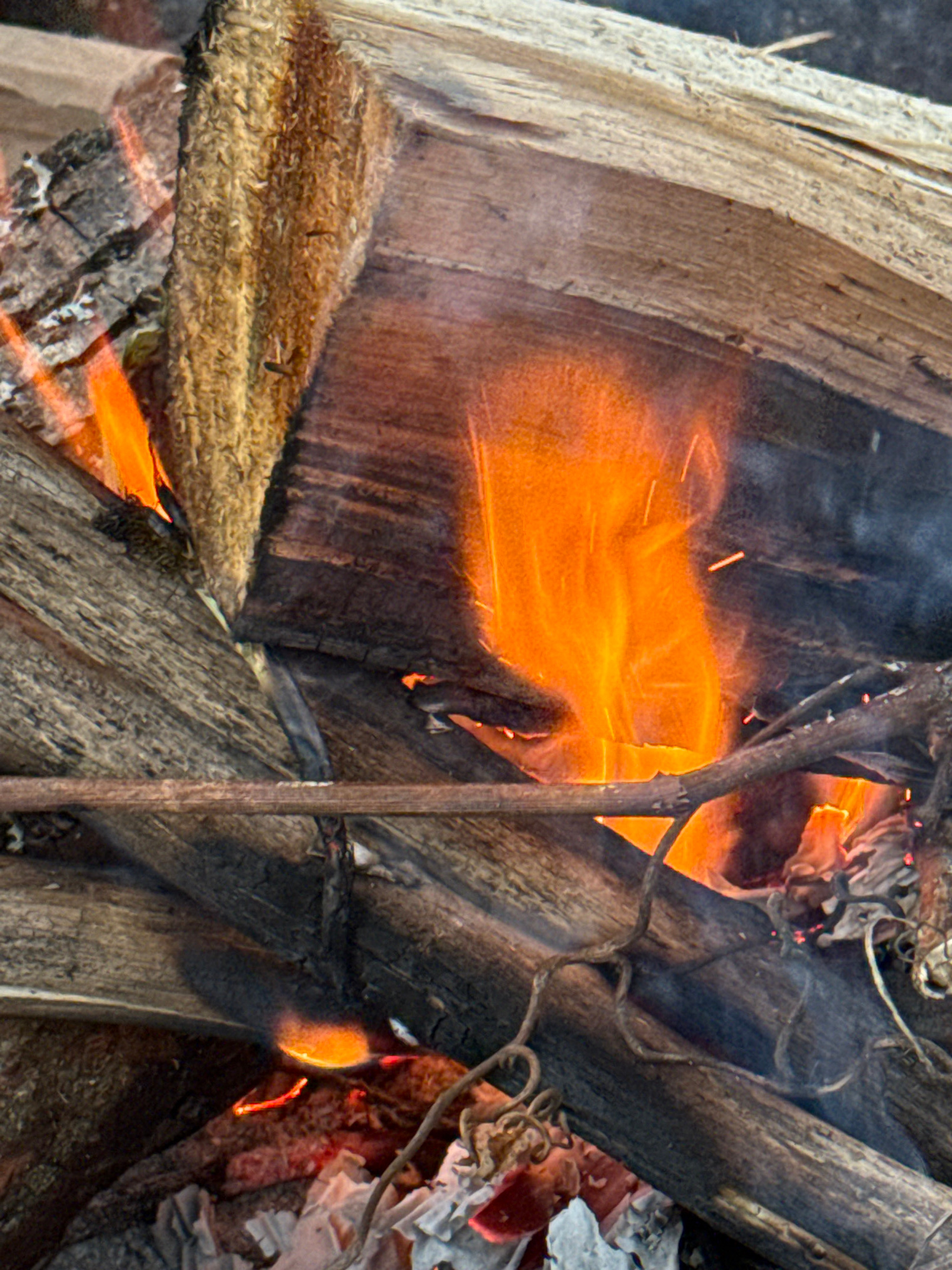 A close-up, angled shot captures a campfire in progress. The fire, characterized by vibrant orange and yellow flames, is central to the image, illuminating the surrounding dry, charred logs. Several pieces of wood are visible, some cracked and weathered, others showing signs of recent burning. Fine tendrils of smoke rise, mingling with the flames. The ground beneath the fire appears to be littered with debris, including what looks like crumpled paper or ash. The overall impression is one of a small, controlled burn in an outdoor, natural setting, likely in the city of Avtomobilist-1 SNT, Russia.