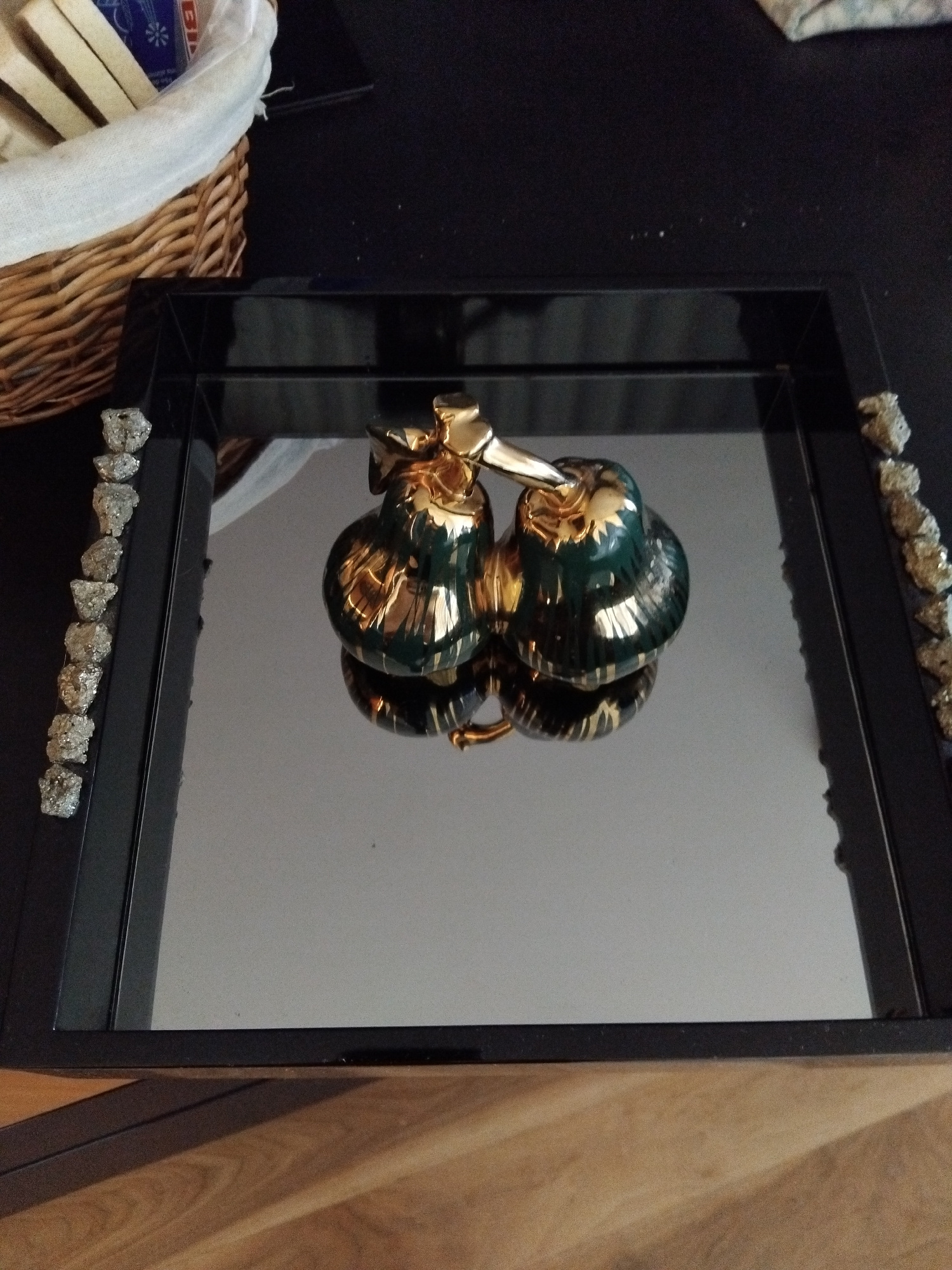 A black rectangular tray with a mirrored surface sits on a wooden surface. On the tray, two decorative green and gold objects resembling pears are placed. The objects are shiny and have a detailed design. Along the left and right edges of the tray, there are rows of small, rough, silver-colored objects that appear to be decorative stones or crystals. In the background, a wicker basket filled with white fabric and some flat, light-colored items is partially visible. The overall scene is dimly lit, with reflections visible on the mirrored surface of the tray.