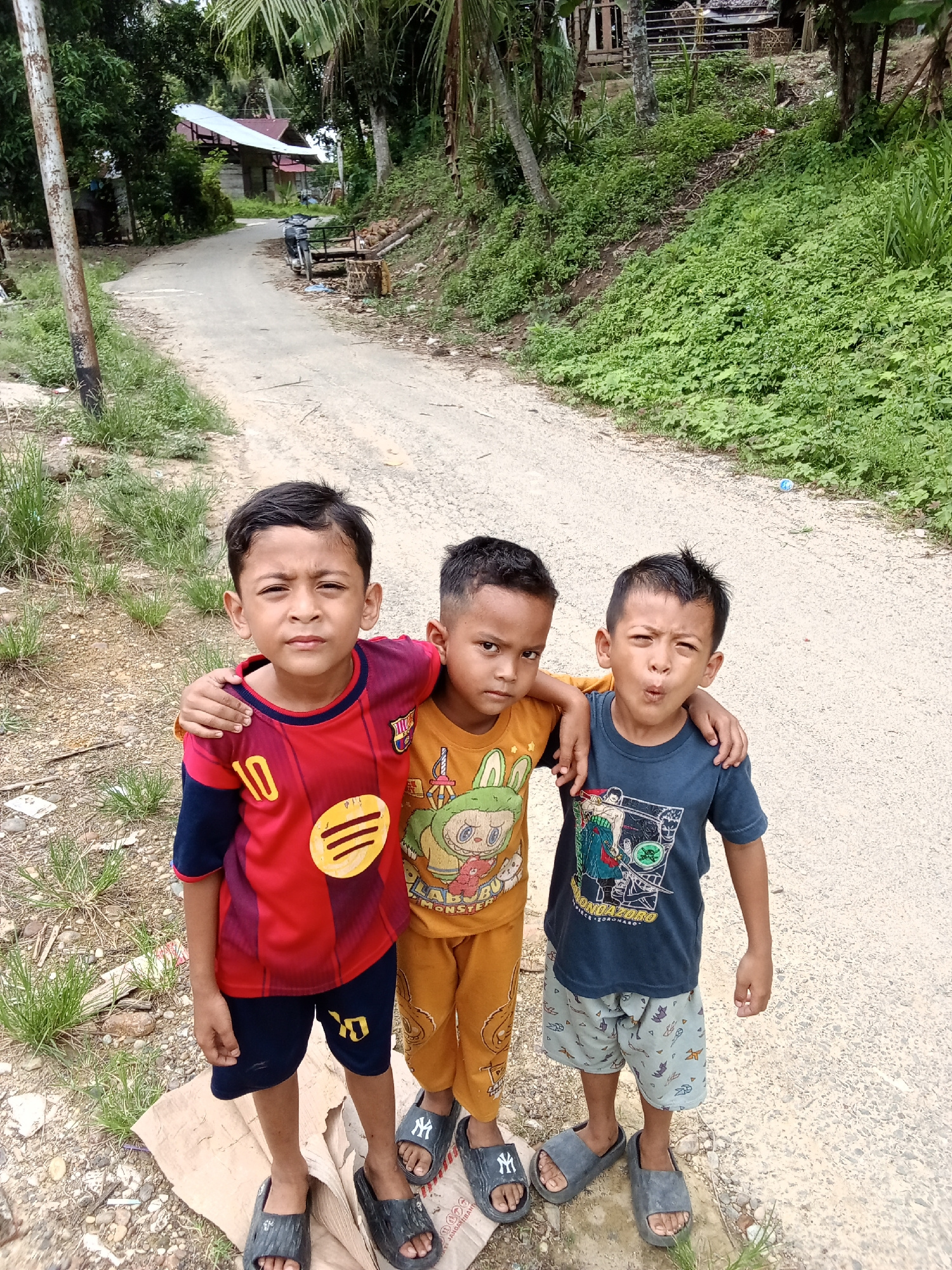 Three young boys stand on a dirt road, posing for a photo. The boy on the left wears a red and blue soccer jersey with the number 10. The middle boy is dressed in an orange t-shirt with a cartoon character and orange pants. The boy on the right wears a dark blue t-shirt with a graphic print and patterned shorts. They are standing with their arms around each other. The road is bordered by lush green vegetation on the right and trees on the left. A small building with a metal roof is visible in the background on the left side of the road. The environment suggests a rural or semi-rural setting. The overall impression is a candid snapshot of children in their local environment.
