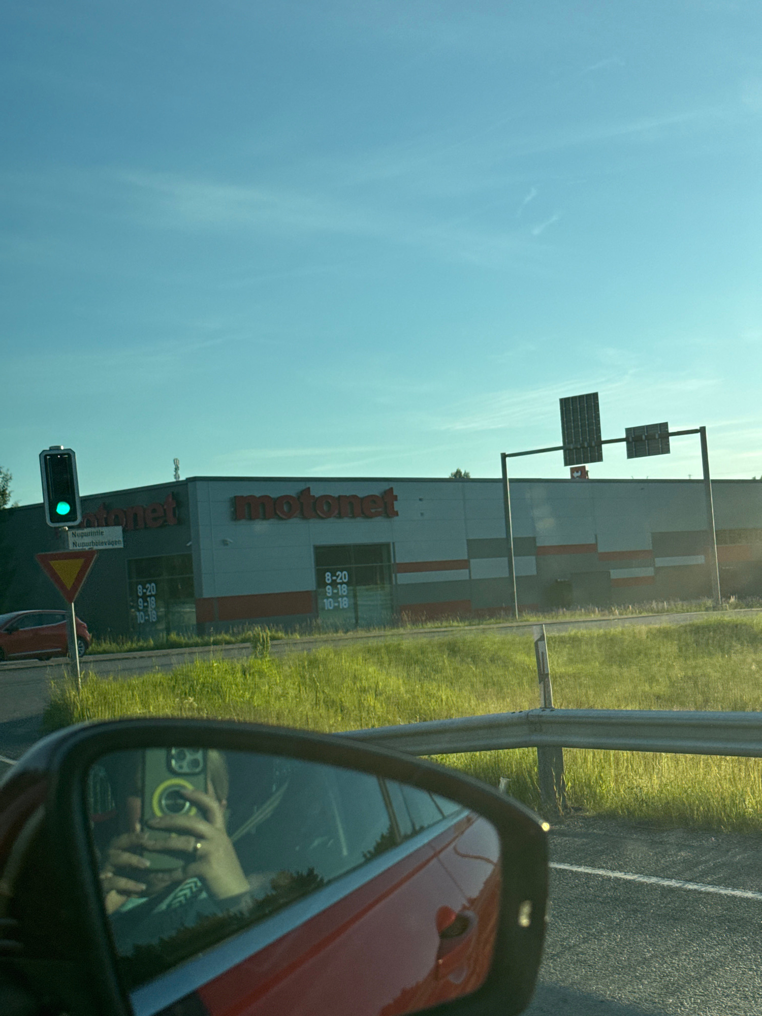 The image captures a view from inside a car, looking out towards a large building with the "motonet" logo. A traffic light showing green is visible on the left, along with a triangular yield sign. The building appears to be a retail store, with operating hours displayed on its facade. The environment is sunny, with green grass and vegetation surrounding the building. A side mirror of the car reflects the driver holding a phone, taking the picture. The sky is clear and blue.