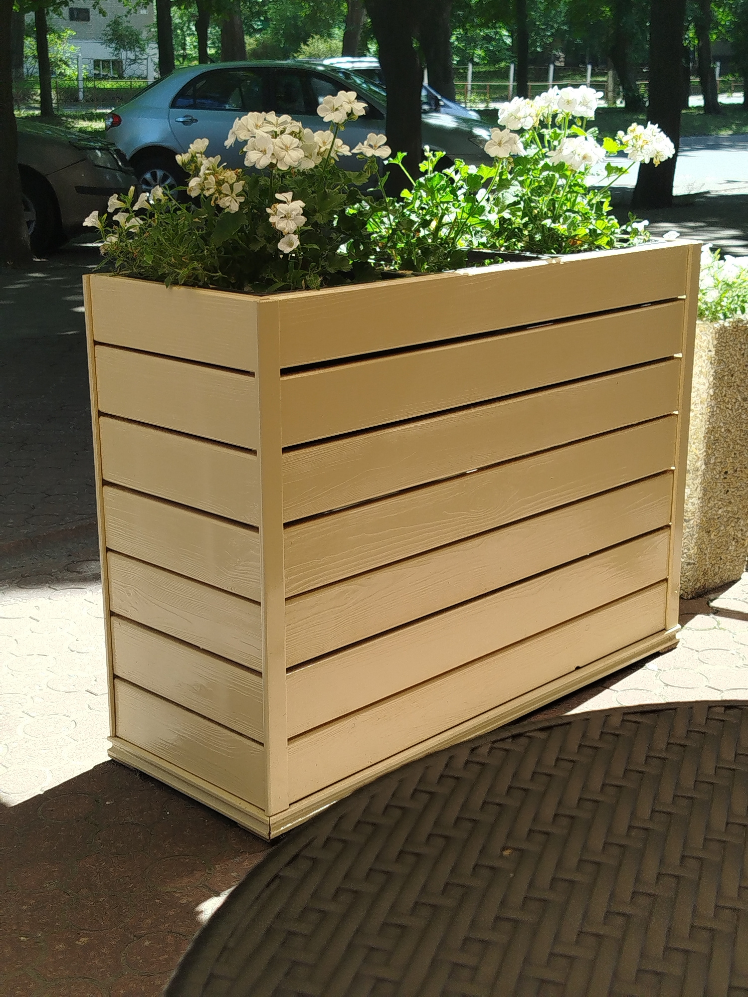 A rectangular, light tan planter box filled with white flowers is positioned outdoors. The planter has a slatted design with horizontal wooden planks. Behind the planter, two cars are visible parked on what appears to be a street or parking area. The environment is characterized by trees casting shadows on the ground, suggesting it is daytime. Part of a dark, woven outdoor table is visible in the foreground, casting a shadow from the planter. The scene is located in Svitlovodsk, Ukraine.