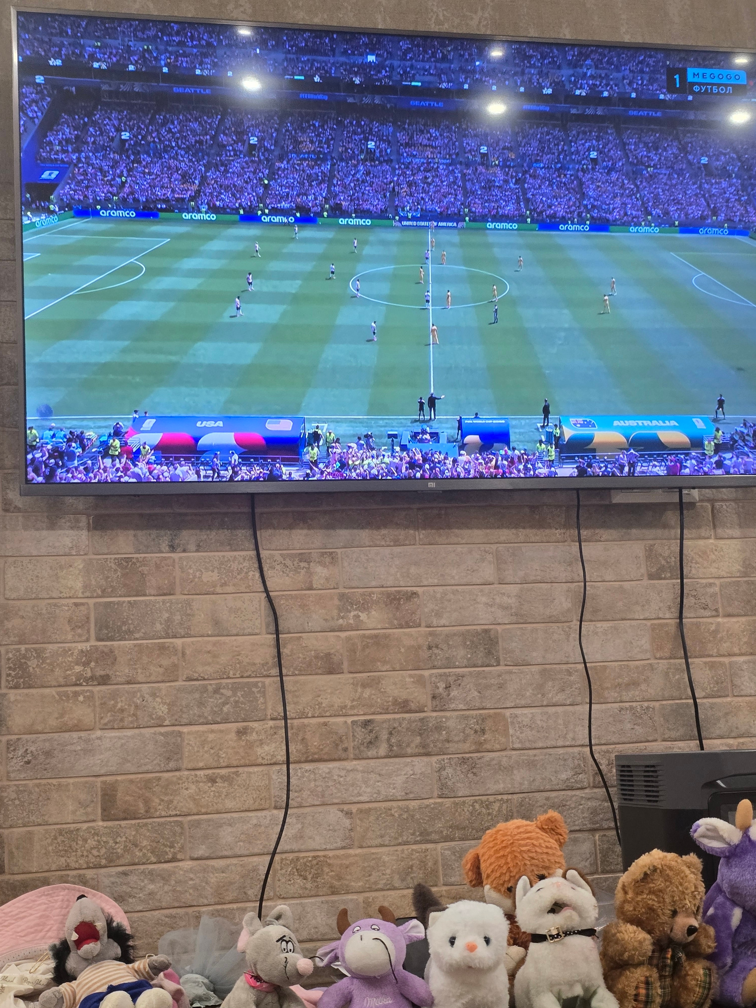 An overhead shot shows a large flat-screen television displaying a soccer match. The stadium is filled with spectators dressed in purple and white. The field is green with white markings. In the foreground, several stuffed animals are arranged on a brick surface. These include a grey mouse, a white cat, an orange fox, a brown bear, and a purple cow. A black cable runs from the television down the brick wall. The scene is indoors.