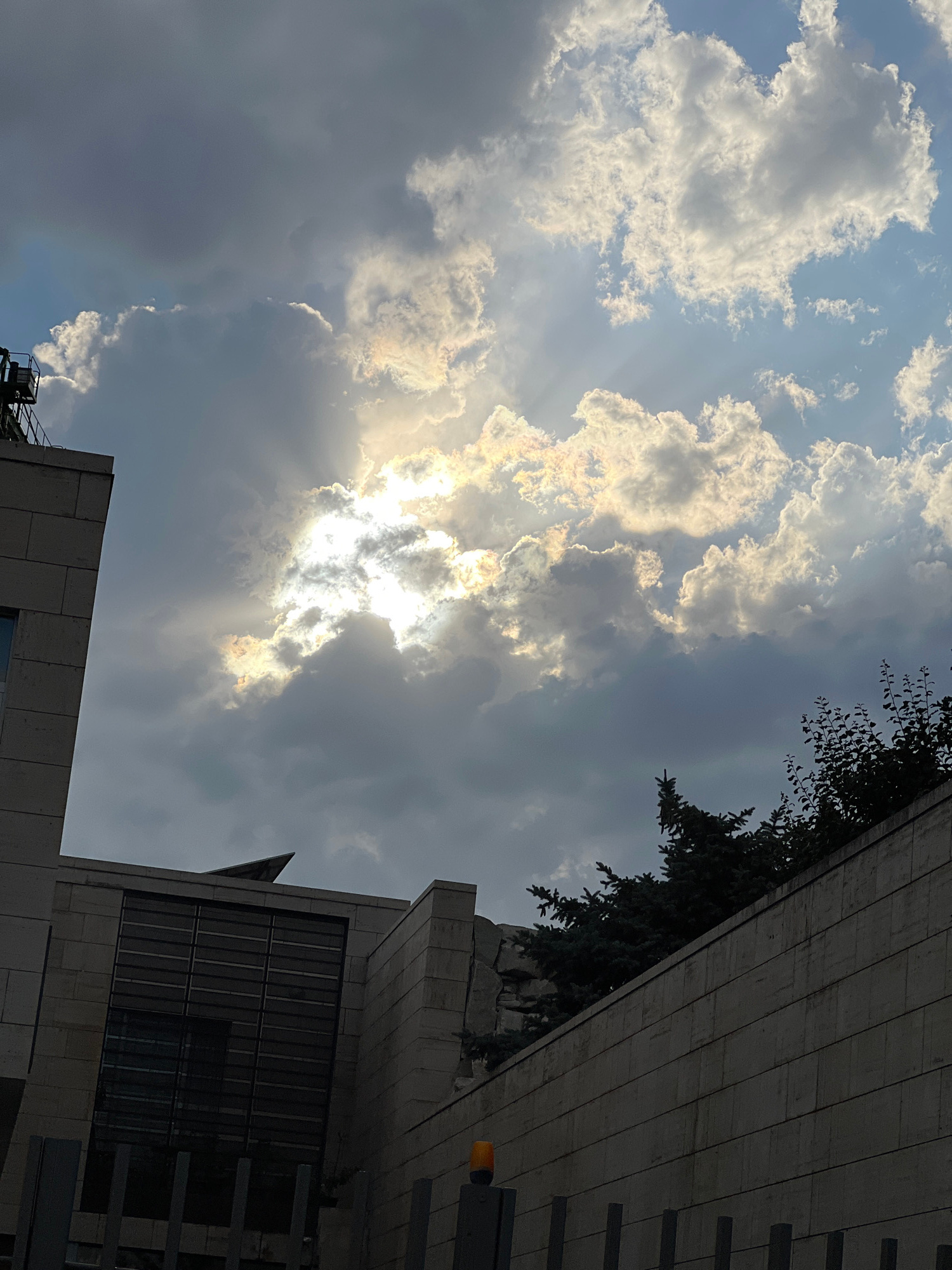 The image shows a dramatic sky with the sun peeking through thick, textured clouds. The sunlight creates bright rays that pierce the gloom, illuminating parts of the clouds with an almost iridescent glow. This creates a sense of awe and wonder.
In the foreground, the angular, modern architecture of buildings is visible, partially obscuring the sky. The buildings are made of concrete and glass, with clean lines and a somewhat imposing presence. A few dark green trees, possibly conifers, are interspersed with the architecture, adding a touch of nature to the urban landscape.
The scene appears to be set in an urban environment, likely Yerevan, Armenia, given the architectural style. The time of day is difficult to pinpoint precisely due to the cloud cover, but the intense sunlight suggests it could be late afternoon or early morning. The weather is cloudy, but with periods of bright sunshine.
No people are visible in the image, and there is no discernible text. The overall mood is one of dramatic natural beauty juxtaposed with man-made structures, evoking a feeling of the sublime.