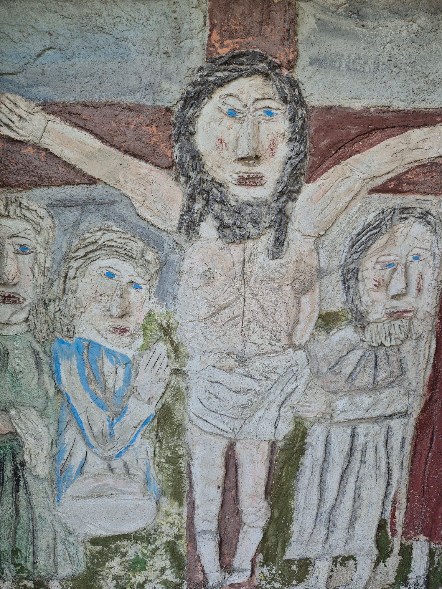 This is a close-up, textured bas-relief sculpture depicting several figures. The style is somewhat crude and stylized, with raised lines and forms carved into a textured surface, possibly concrete or stucco. The colors are muted, with earthy tones of brown, beige, and grey, accented by patches of blue and some green.
The primary subject appears to be a crucifixion scene or a related religious motif. The central figure, positioned horizontally, has arms outstretched and is depicted with a pale body, dark hair, and prominent blue eyes. The body is wrapped in a rough, textured cloth. To the left of this figure, an arm is extended downwards, with a hand showing carved fingers.
Above the central figure, another face is visible, also with blue eyes, a beard, and what seems to be a robe or cloak. Below the central figure, two more figures are partially visible. One appears to be lying down, with a face turned upwards and blue eyes. Another figure, further down and to the right, is also present with visible facial features. The overall arrangement suggests a narrative scene, possibly a depiction of suffering or lamentation, common in religious art.
The textured surface, with its cracks and rough patches, gives the artwork a weathered and ancient feel. The limited color palette and the abstract, almost primitive rendering of the figures contribute to a solemn and impactful visual. There is no visible text in the image. The location context is Rottendorf, Germany, which may indicate the artwork is part of a church, cemetery, or public art installation in that region. The time of day and weather are not discernible from the static image.