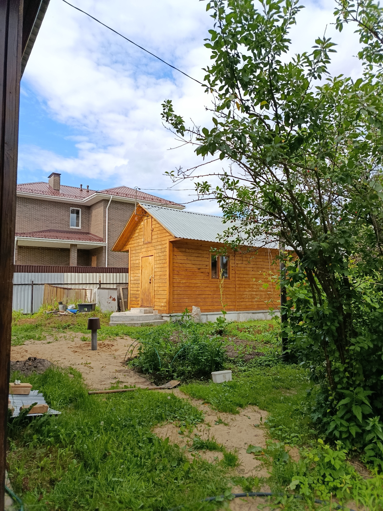 This image depicts a backyard scene on a cloudy day. In the foreground, a path made of dirt and gravel winds through lush green grass and weeds. To the left, a wooden structure, possibly a fence or part of a building, frames the view. A modern brick house with a red-tiled roof stands in the background, partially obscured by a smaller wooden shed. The shed has a metal roof and a wooden door, and sits on a concrete foundation.
Various objects are scattered around the yard, including a metal post with a cap, some construction materials like bricks and metal sheeting, a blue bucket, and a black tub. Overgrown vegetation, including a large leafy tree on the right side of the frame, suggests the area is not meticulously maintained. The sky is filled with white clouds against a light blue background. The overall impression is of a rural or suburban backyard under development or undergoing natural overgrowth, with a sense of a quiet, ordinary day. There is no visible text in the image.