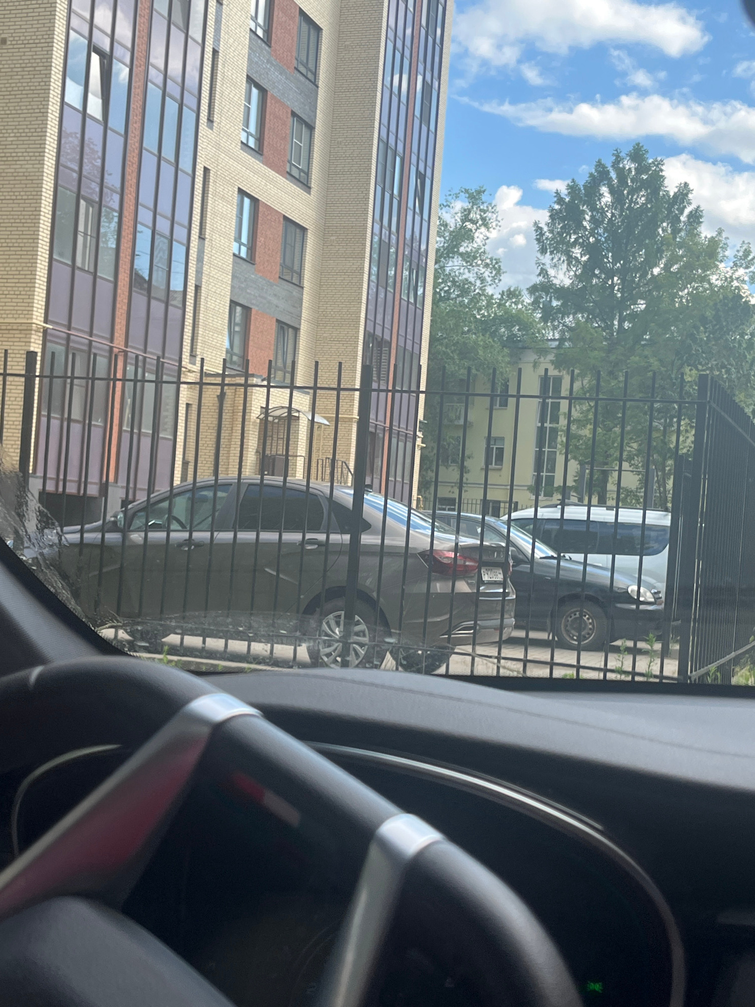 This is a photograph taken from inside a vehicle, looking out through the windshield and past a black metal fence. The scene depicts a residential area in Kostroma, Russia.
In the foreground, a portion of the car's steering wheel is visible on the right side of the frame, along with parts of the dashboard. The view through the windshield is dominated by a multi-story brick building with numerous windows, some of which reflect the blue sky and white clouds. Adjacent to this building, a dark-colored car is parked, partially obscured by the fence. Further in the background, more trees and another building, painted a pale yellow, can be seen.
The lighting suggests it is daytime, likely late morning or afternoon, with bright sunlight casting reflections. The sky is partly cloudy. The presence of a fence and parked cars indicates a typical urban or suburban residential setting. No people are clearly visible in the frame. There is no discernible text in the image.