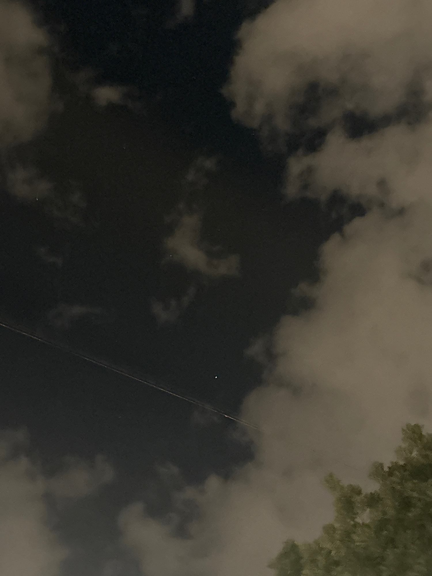 The image shows a night sky with clouds and a distant light source, possibly a star or a planet. A faint line, possibly a contrail or wire, stretches diagonally across the sky. Part of a tree is visible in the lower right corner. The image does not contain any identifiable landmarks or elements that would allow for the determination of its location in Plovdiv, Bulgaria.