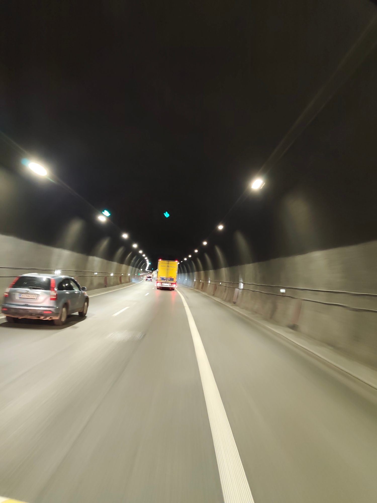 The image captures a forward-looking perspective from within a highway tunnel. The tunnel is illuminated by rows of lights mounted on the ceiling, casting a bright glow on the roadway. The walls of the tunnel appear to be made of concrete. Several vehicles are visible on the road: a silver SUV is in the left lane, and further ahead, a yellow truck with its brake lights illuminated is in the right lane. The road is marked with a white line separating the lanes, and there are some traffic lights visible, showing green signals. The overall impression is one of movement and transit through an enclosed passage. The location is identified as being in the city of Gorno Kamartsi, Bulgaria.
