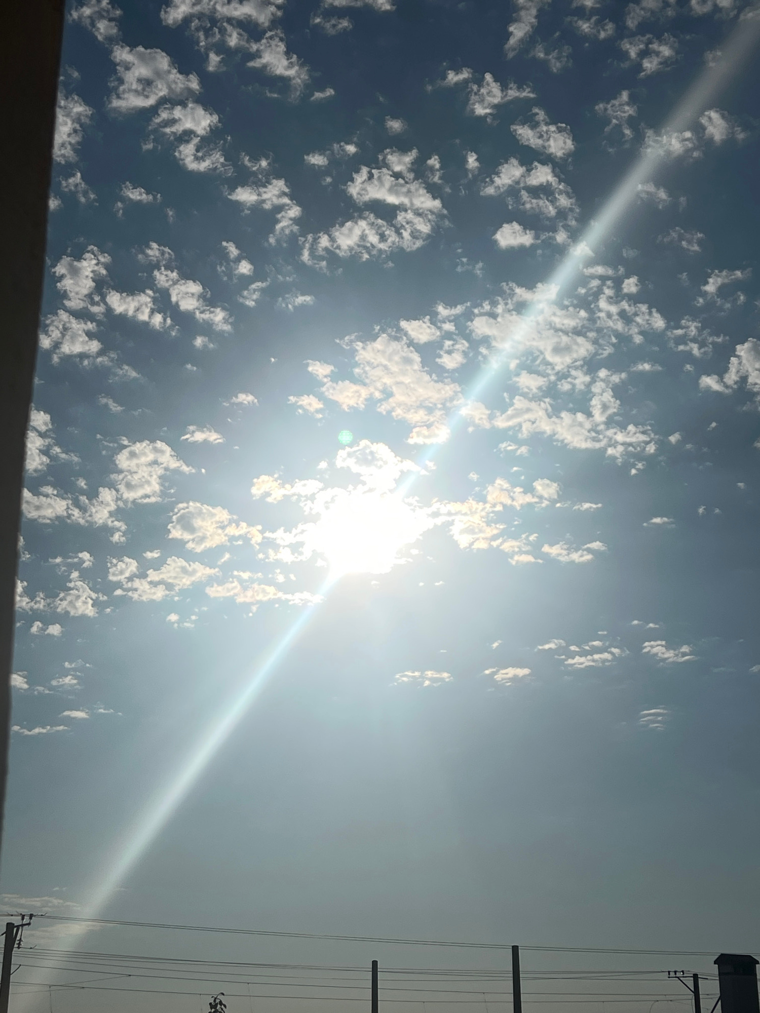 The image captures a bright, sunlit sky filled with scattered cumulus clouds. The sun is positioned in the upper central portion of the frame, partially obscured by clouds, and emits a strong glare that creates a prominent light streak extending diagonally across the sky from the lower left to the upper right. The sky transitions from a deep blue at the top to a lighter, hazy blue towards the bottom. In the lower section of the image, silhouetted against the bright sky, are power lines supported by poles, and what appears to be a small building or structure on the far right. The scene is devoid of any people or discernible activity. The setting is not specifically identifiable as Sochi, Russia, based on the visual information provided.