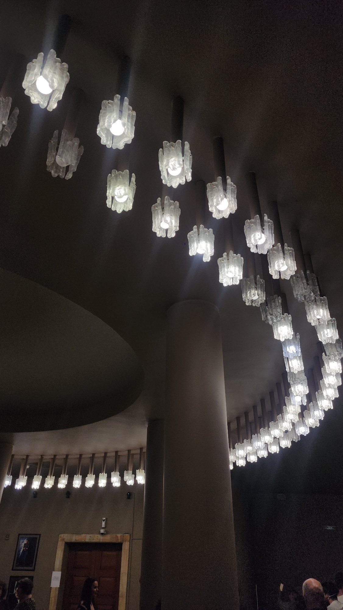 This image depicts an interior space with a distinctive ceiling adorned with multiple rows of art deco style chandeliers. The chandeliers are made of clear, textured glass pieces that surround individual light bulbs, creating a diffused glow. They are suspended from the dark ceiling in a curved arrangement, extending across a significant portion of the visible space.
In the lower part of the image, architectural elements such as a large, rounded column and a textured wall are visible. On the wall, there is a framed picture and a wooden doorway. A few individuals are present in the lower section, including two women in the foreground, one looking towards the camera. Another person, a man with a bald head, is partially visible in the bottom right corner. The lighting is predominantly from the chandeliers, casting a warm, ambient light, with shadows present due to the dim overall illumination.
The scene is identified as being inside the State Kremlin Palace in Moscow, Russia. The palace is known for its impressive architecture and is used for various events, including concerts and official receptions. The chandeliers are a notable design feature within the building's interiors, contributing to its opulent atmosphere.