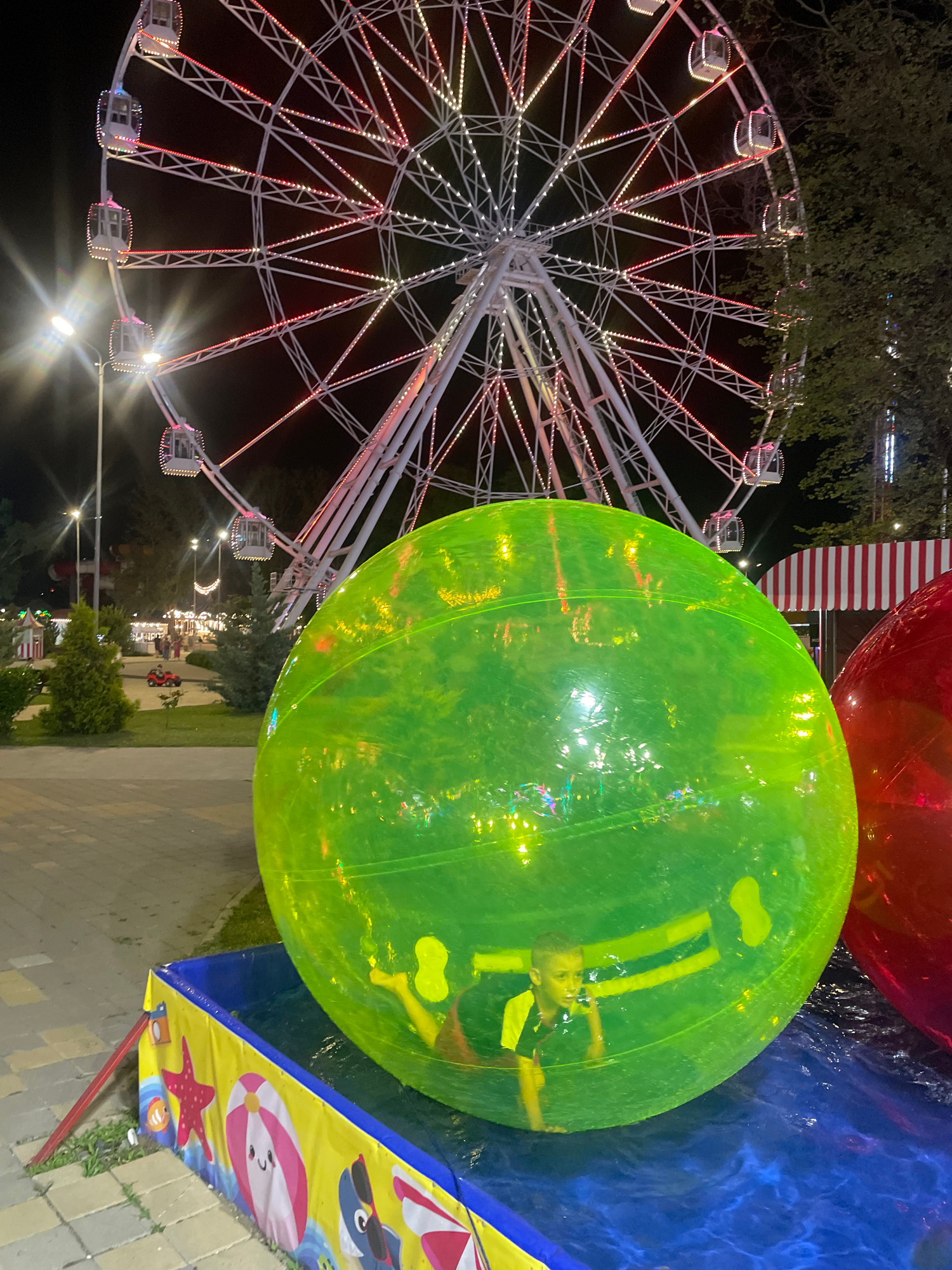 An eye-level, full shot captures a boy inside a large, translucent green sphere, illuminated by a Ferris wheel at night in an amusement park. The boy, appearing to be in his early elementary school years, wears a dark shirt with yellow accents and is positioned with his back against the sphere's interior, looking forward with a determined expression. The sphere, filled with air, rests in a blue pool of water, with a portion of a red sphere visible to the right. The Ferris wheel, adorned with white lights and pink and yellow accents, dominates the background, with its structure and cabins clearly visible against the dark night sky. Streetlights with bright white beams are also present, along with trees and park structures in the mid-ground. The scene is likely in a public amusement park or entertainment venue.