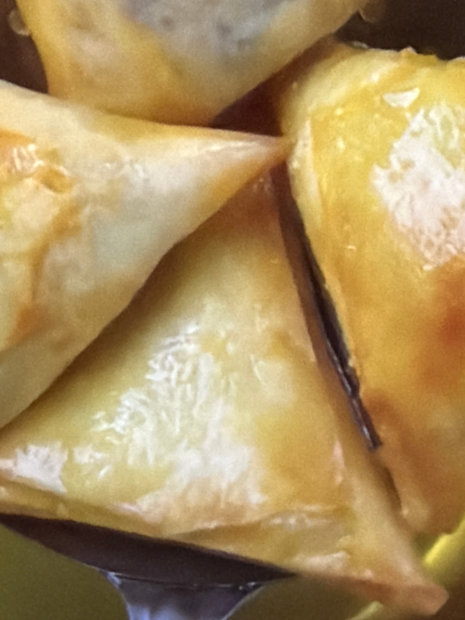A close-up, top-down shot captures several triangular pastries with golden-brown, flaky wrappers. The pastries are layered and slightly overlapping, suggesting they are part of a larger dish. One pastry in the foreground shows a dark filling peeking through a slight opening in its wrapper, hinting at a sweet or savory interior. The texture of the wrappers appears crisp and slightly oily, with some areas exhibiting a glossy sheen. The lighting creates highlights and shadows, emphasizing the dimensionality of the pastries. The overall impression is one of freshly baked, enticing food.
