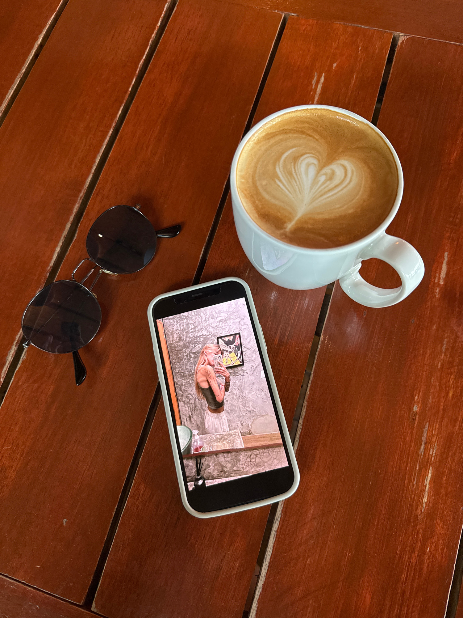 On a warm, reddish-brown wooden table, likely outdoors in Rawai, Thailand, sit a white ceramic coffee cup filled with latte art, a pair of dark-lensed, round sunglasses, and a smartphone displaying a selfie. The coffee cup, with a heart design in the foam, is positioned to the right of the frame. The sunglasses are to the left, with their arms extended. The smartphone is in the foreground, its screen showing a person with blonde hair taking a mirror selfie in what appears to be a bathroom. The table's surface is made of parallel wooden planks, showing some wear and tear.