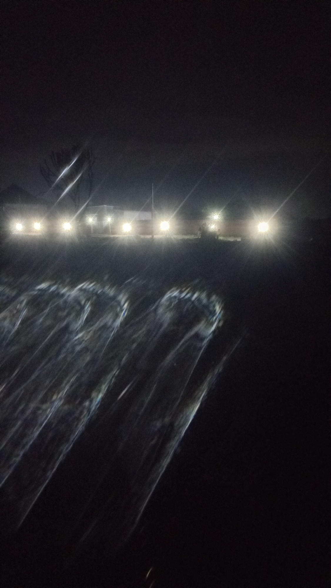 The image depicts a nighttime scene with multiple bright headlights illuminating the foreground, creating streaks of light across a dark surface that appears to be wet or reflective, possibly due to rain or puddles. In the background, several vehicles are visible, their headlights also shining brightly, suggesting they are on a road or in a parking area. A few indistinct structures, possibly buildings, are located behind the vehicles. The overall atmosphere is dark and somewhat hazy, with the light sources being the most prominent features. The location is identified as Kwang, Nigeria, however, specific details of this city are not discernible from the image. There are no clear individuals or specific actions being depicted.