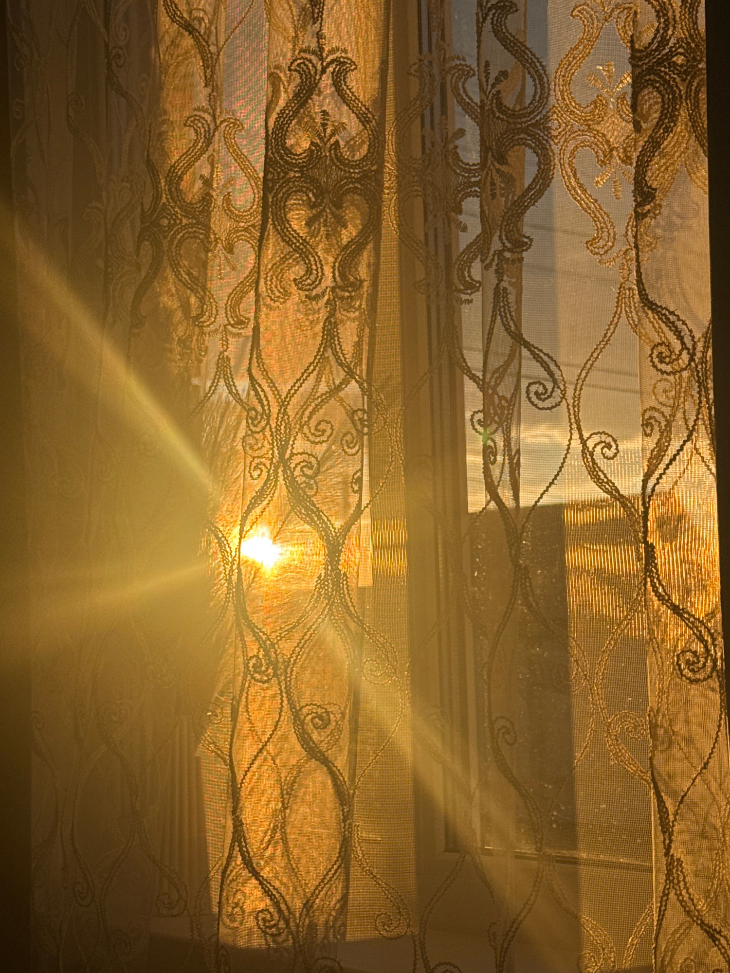 The image depicts a sunbeam, likely from a setting or rising sun, piercing through a decorative sheer curtain. The curtain has an ornate, scrolled pattern in a darker thread. The light creates a strong lens flare effect and illuminates the fabric, casting shadows of the pattern onto itself and the unseen window and background. The overall color palette is dominated by warm golden and orange hues. The scene appears to be an interior view looking outwards through a window. There are no people or discernible objects other than the curtain and the light. The specific location in the city of Rayevskiy, Russia, cannot be determined from the visual information provided in the image.