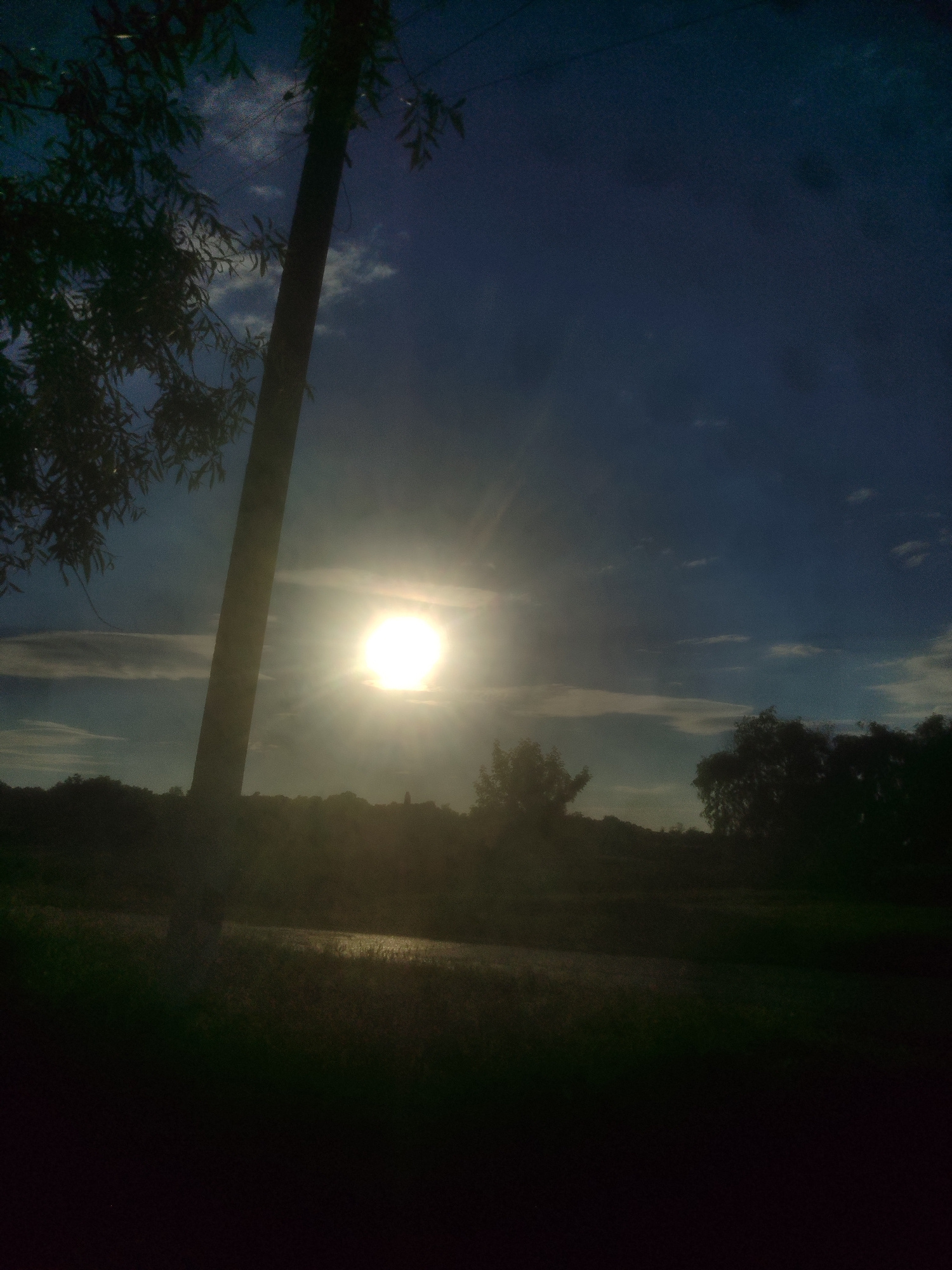 The image captures a silhouetted landscape under a dramatic sky. A tall, dark pole stands prominently in the left foreground, partially obscuring a bright, luminous sun that dominates the center of the frame. The sun is surrounded by a halo of light, indicating it is low in the sky, suggesting either sunrise or sunset. Wisps of clouds are visible, illuminated by the sun's intense glow, and contrast with the deep blue of the upper sky.
The middle ground features a dark, uneven line of trees and vegetation silhouetted against the bright horizon. Below the trees, a body of water or a damp field reflects the sunlight, creating a shimmering effect. The foreground is almost entirely black, suggesting a very dark, possibly grassy area.
The overall impression is one of natural beauty, with the powerful light of the sun creating a striking visual. The atmosphere is serene, with a hint of mystery due to the silhouetted elements. There is no visible text in the image. The time of day is clearly either dawn or dusk, and the weather appears clear apart from the clouds. The location is likely outdoors, possibly a rural or natural setting with a body of water.