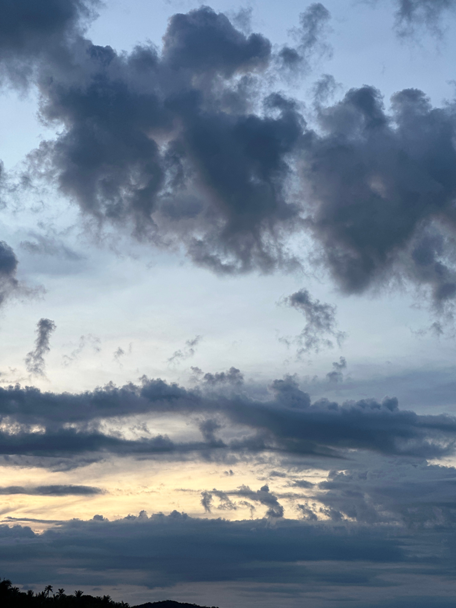 The image shows a dramatic sky filled with dark, brooding clouds. The lighting suggests either sunrise or sunset, with a sliver of pale yellow light peeking through the clouds near the horizon. In the foreground, the silhouetted outline of a tropical coastline is visible, with palm trees and undulating hills. The location is identified as Baan Mae Nam, Thailand. There are no people or active subjects visible in the image; it is a landscape focusing on the atmospheric conditions.