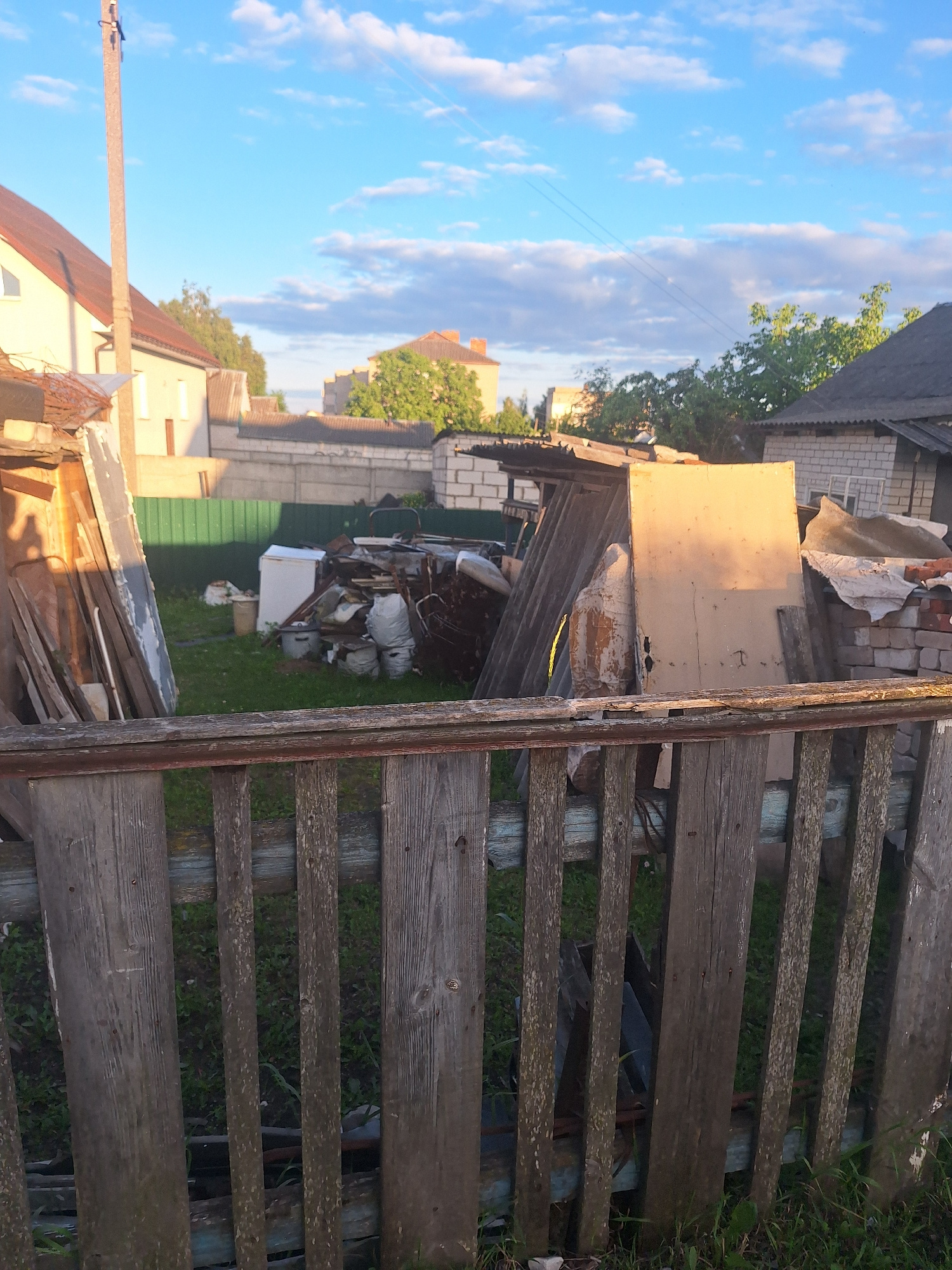 This image depicts a cluttered backyard in Kapyĺ, Belarus, under a partly cloudy blue sky. The foreground is dominated by a weathered wooden fence with vertical planks, some showing signs of decay and blue paint. Beyond the fence, a grassy area is filled with various discarded items. These include stacks of lumber, sheets of corrugated metal or similar material, several bags that appear to contain debris or soil, buckets, and a white refrigerator. In the background, there are residential buildings with visible roofs and walls, as well as trees and a green fence. The overall impression is one of disarray and storage of old materials.