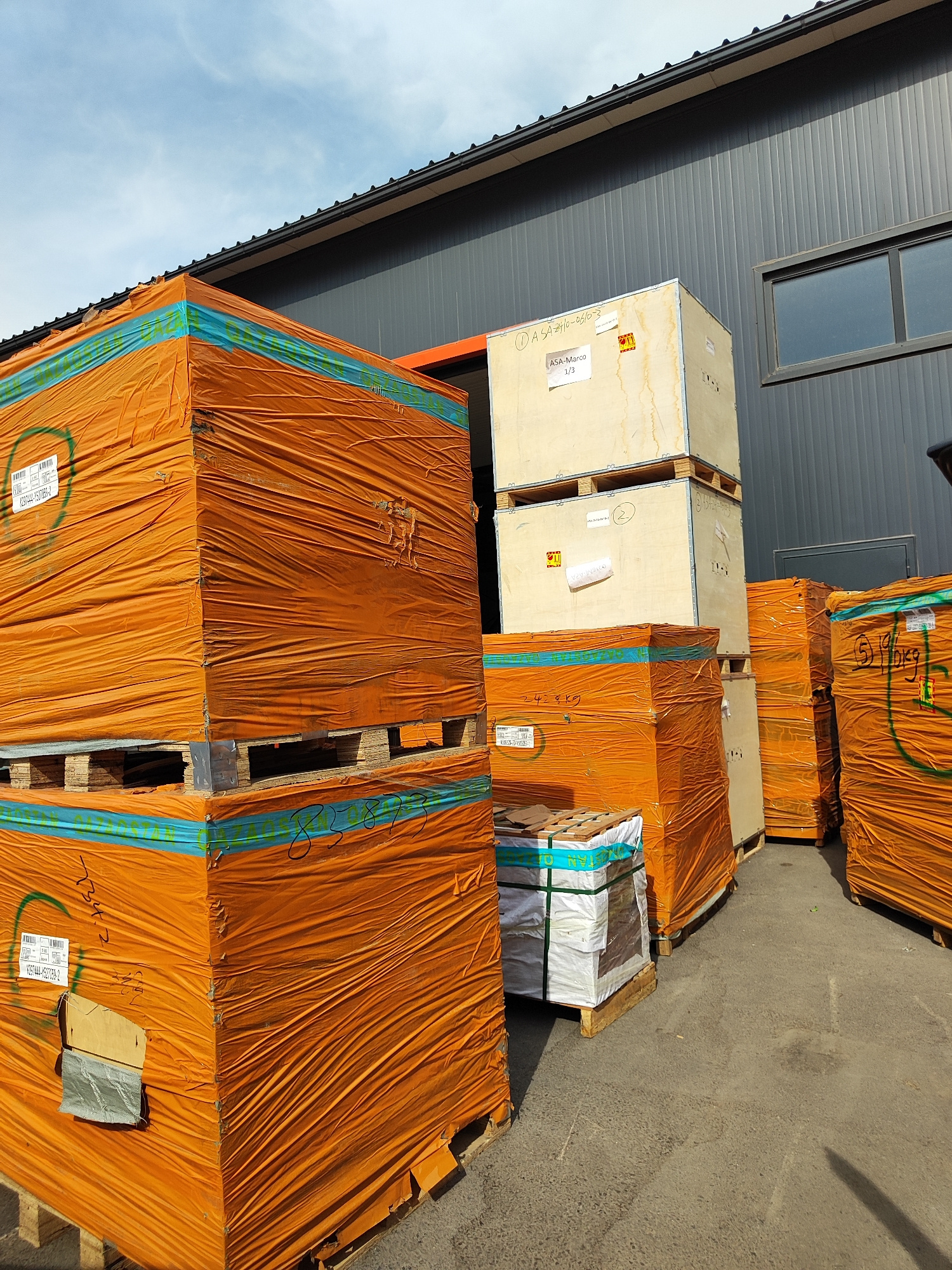 This media file depicts an outdoor storage area, likely a loading dock or warehouse exterior, in Boralday, Kazakhstan. The scene is dominated by large, stacked pallets of goods wrapped in bright orange plastic sheeting. The pallets are of varying sizes and appear to be made of wood. Some of the stacks are very tall, suggesting significant quantities of material.
In the background, a modern-looking industrial building with a dark gray, ribbed facade and a black roof is visible. A window with a reflective surface is present on the building. The sky above is partly cloudy with a light blue hue, indicating daytime.
Notable details include various labels and markings on the wrapped pallets and wooden crates. Some visible text on a light-colored wooden crate reads "ASA-Marco 1/3" and "ASA2410-06/07-3". Another crate has a label with what appears to be a barcode and more text. Some of the orange wrapping also has blue banding with handwritten numbers, such as "85873". One smaller pallet in the foreground contains items wrapped in white and clear plastic.
The overall impression is one of a commercial or industrial setting where goods are being stored or prepared for shipment. The bright orange wrapping stands out against the muted tones of the building and the asphalt ground. The lighting suggests it is a sunny day. No people are visible in the frame. The stacked nature of the goods and the presence of the industrial building strongly suggest a logistical or warehousing context.