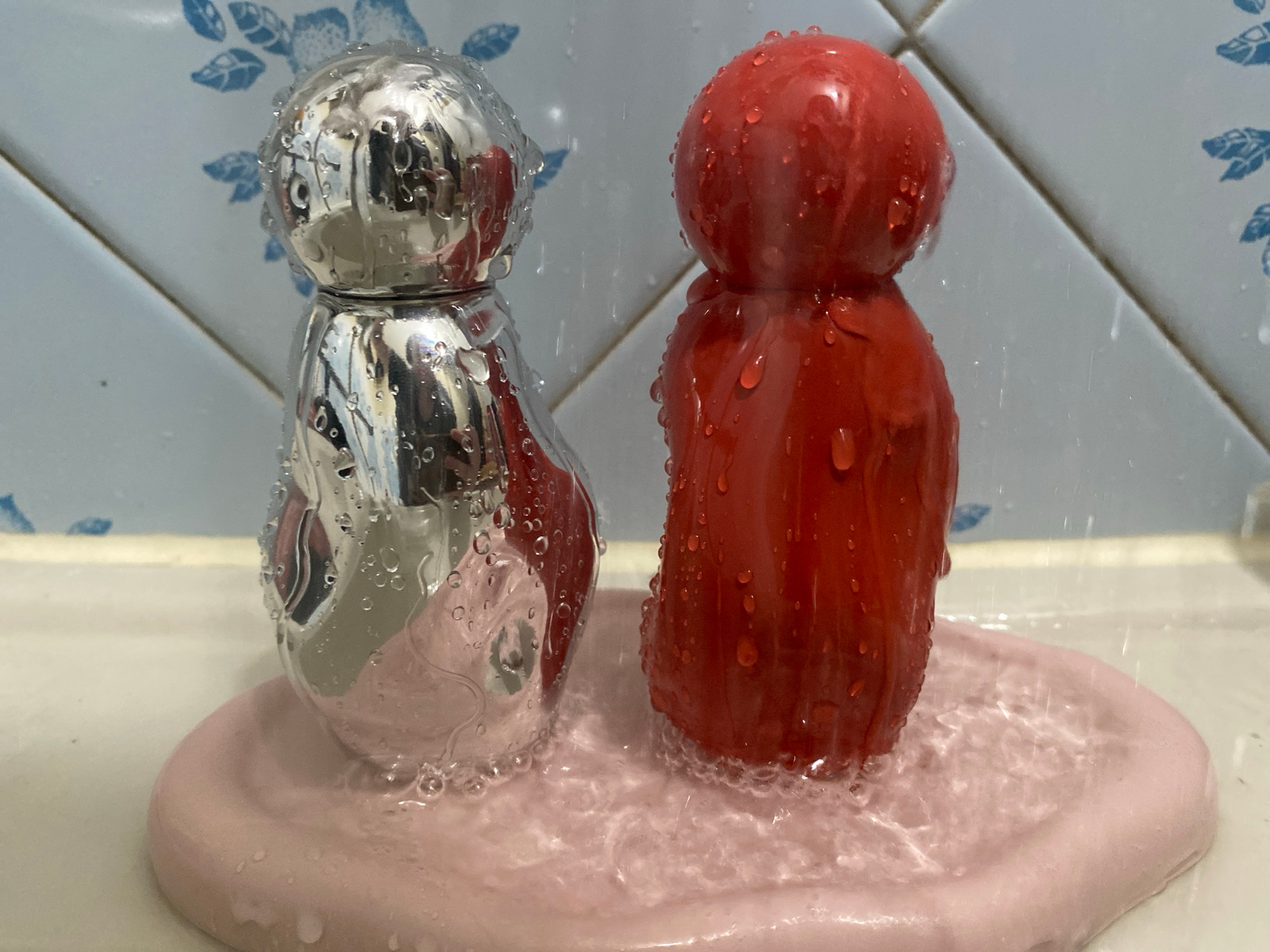 A close-up, eye-level shot captures two abstract, figurine-like objects being rinsed with water. The object on the left is metallic and reflective, with water droplets clinging to its surface. It has a rounded top that tapers down into a wider base. To its right is a similar figurine, this one solid red and textured with numerous water droplets. It also features a rounded top and a tapering lower body. Both objects stand on a pale pink, circular tray that is submerged in water, creating a bubbly, rippling effect around their bases. Behind the figurines, a tiled surface with a blue floral pattern is visible. Water is seen falling from above, indicated by streaks and splashes. The overall setting appears to be a bathroom sink or countertop.