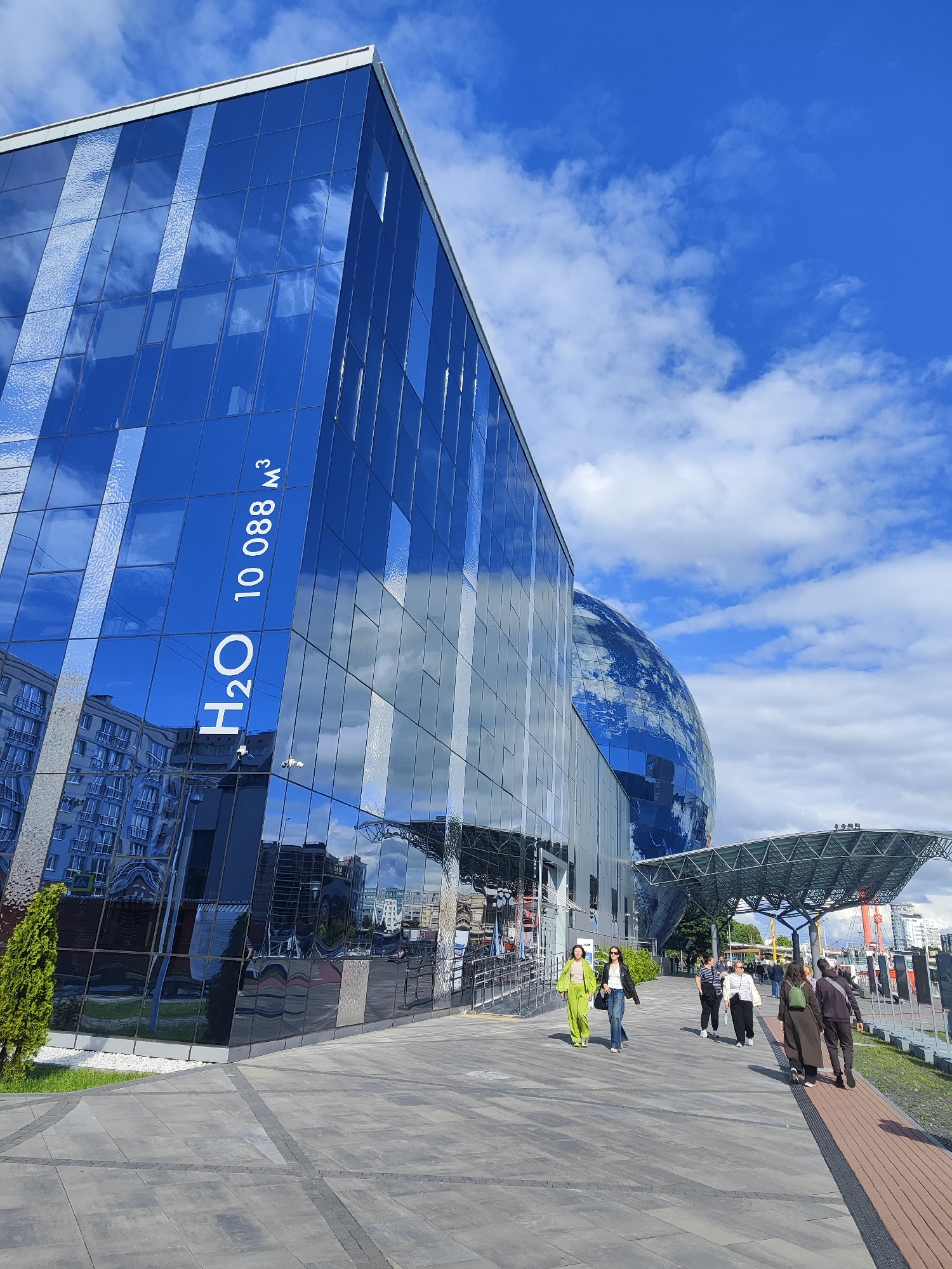 In Kaliningrad, Russia, the facade of a modern building, possibly the "Oceanarium," is predominantly blue glass, reflecting the sky and clouds. A large spherical structure is also visible, also clad in reflective blue glass, possibly part of the same complex. Text reading "H2O 10 088 M³" is prominently displayed in white lettering on the building's glass surface. Several individuals are walking on a paved walkway in front of the building. The sky is bright blue with scattered white clouds. The scene suggests a public space, possibly a tourist attraction or an educational facility.