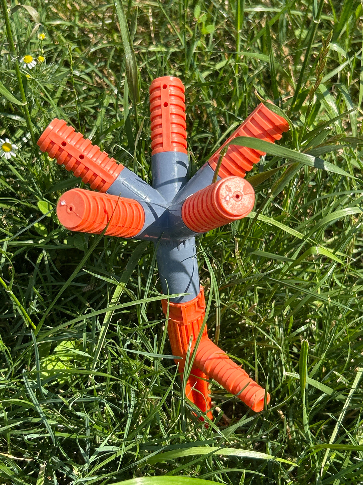 The image shows an orange and grey garden sprinkler lying on the ground amidst lush green grass and a few small white wildflowers. The sprinkler is star-shaped, with multiple arms extending outwards. Each arm is orange and ribbed, with small circular openings near the tip. The central hub of the sprinkler is grey. The grass is tall and dense, with some blades casting shadows, suggesting it is a sunny day. The overall setting appears to be a garden or a grassy field in a rural location. There is no text visible in the image. The scene is peaceful and appears to be outdoors during daylight hours. The sprinkler is not actively in use; it is lying on its side. There are no people or animals present in the frame. The focus is on the garden sprinkler and its natural surroundings. The image evokes a sense of summer or late spring due to the vibrant green grass and blooming flowers. The lighting suggests it could be late morning or early afternoon.
