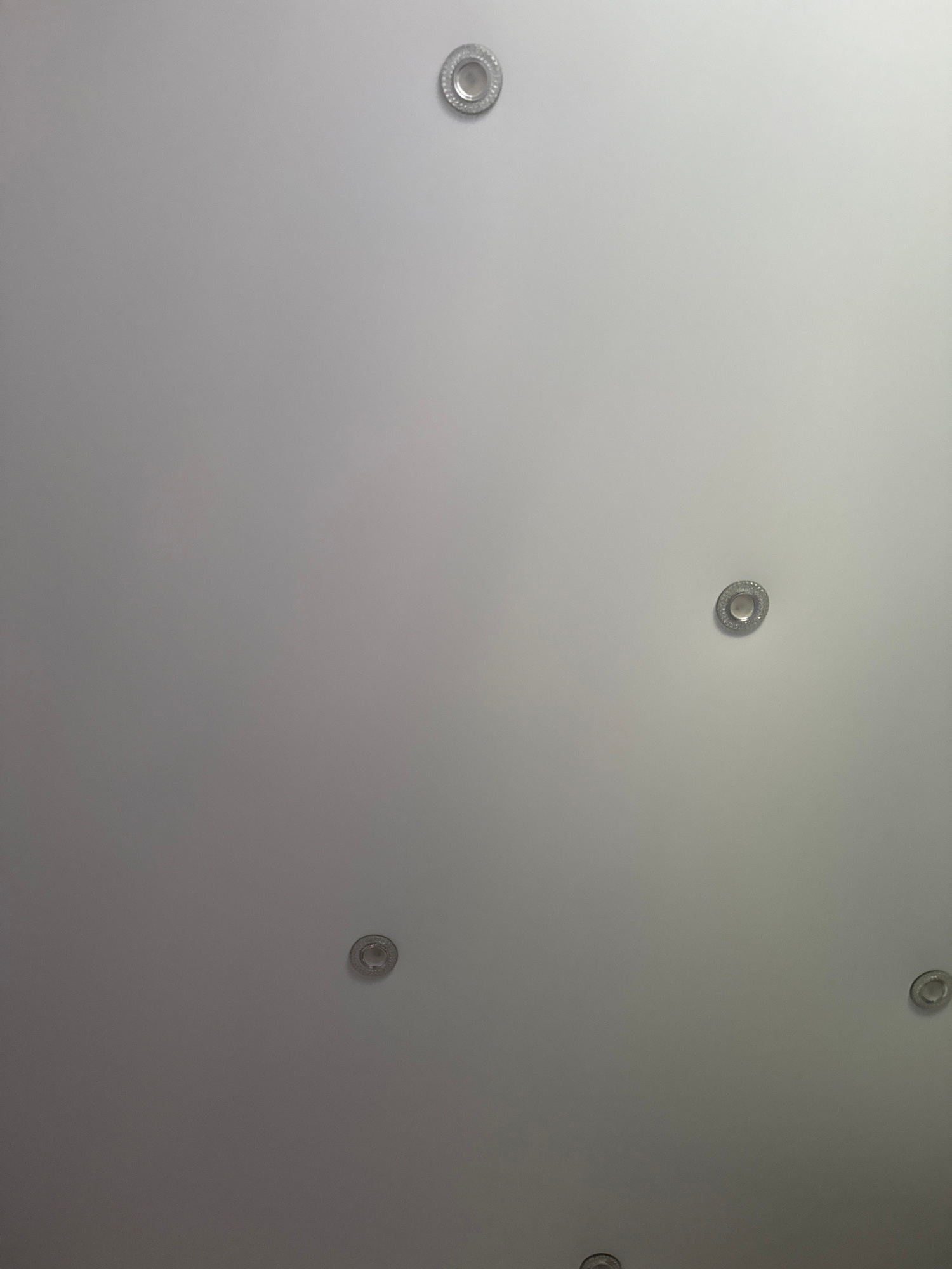 The image shows a section of a plain, light-colored ceiling with several small, circular light fixtures. These fixtures appear to be recessed and have a metallic rim. There are at least five visible fixtures distributed across the ceiling. The lighting conditions are even, suggesting an indoor environment. The scene is devoid of any individuals or discernible objects other than the light fixtures. The image does not provide any information about its location in the city of Asha, Russia, or any context beyond what is visually present.