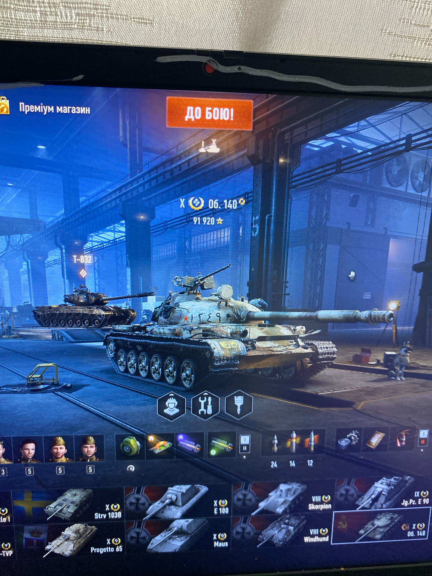 The image shows a computer screen displaying a video game interface, likely a tank simulation game. In the center of the screen, a large battle tank is prominently featured in a dimly lit indoor environment resembling a garage or factory. Other tanks and game-related icons are visible on the screen. The top portion of the screen displays text in Russian, "Преміум магазин" (Premium Store) and "ДО БОЮ!" (To Battle!), indicating game menus or options. There are also numerical indicators and symbols commonly found in game interfaces, such as "X 06. 140" and "91 920".
The image is not of the city of Szczecin, Poland. It is a screenshot of a video game.