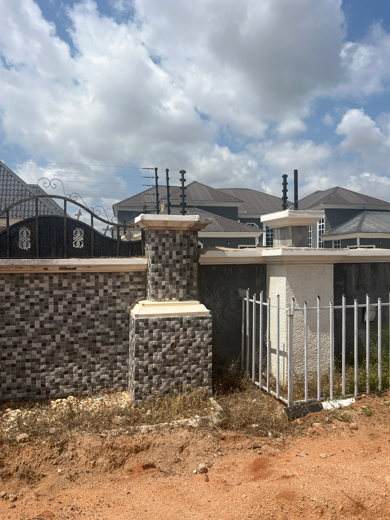 The image depicts a residential property in Howayi, Nigeria, under a cloudy sky. The scene focuses on the exterior of a multi-story house, characterized by its grey tiled roof, white textured walls, and dark-tinted windows. A prominent feature is the dark metal gate adorned with decorative scrollwork, leading to a tiled walkway with a mosaic pattern. Adjacent to the gate is a fence made of white metal bars. The ground is a mixture of red dirt and sparse, dry vegetation. Electrical wires are visible running above the gate and fence, and there are also security spikes installed on top of a wall. The lighting suggests it is daytime.