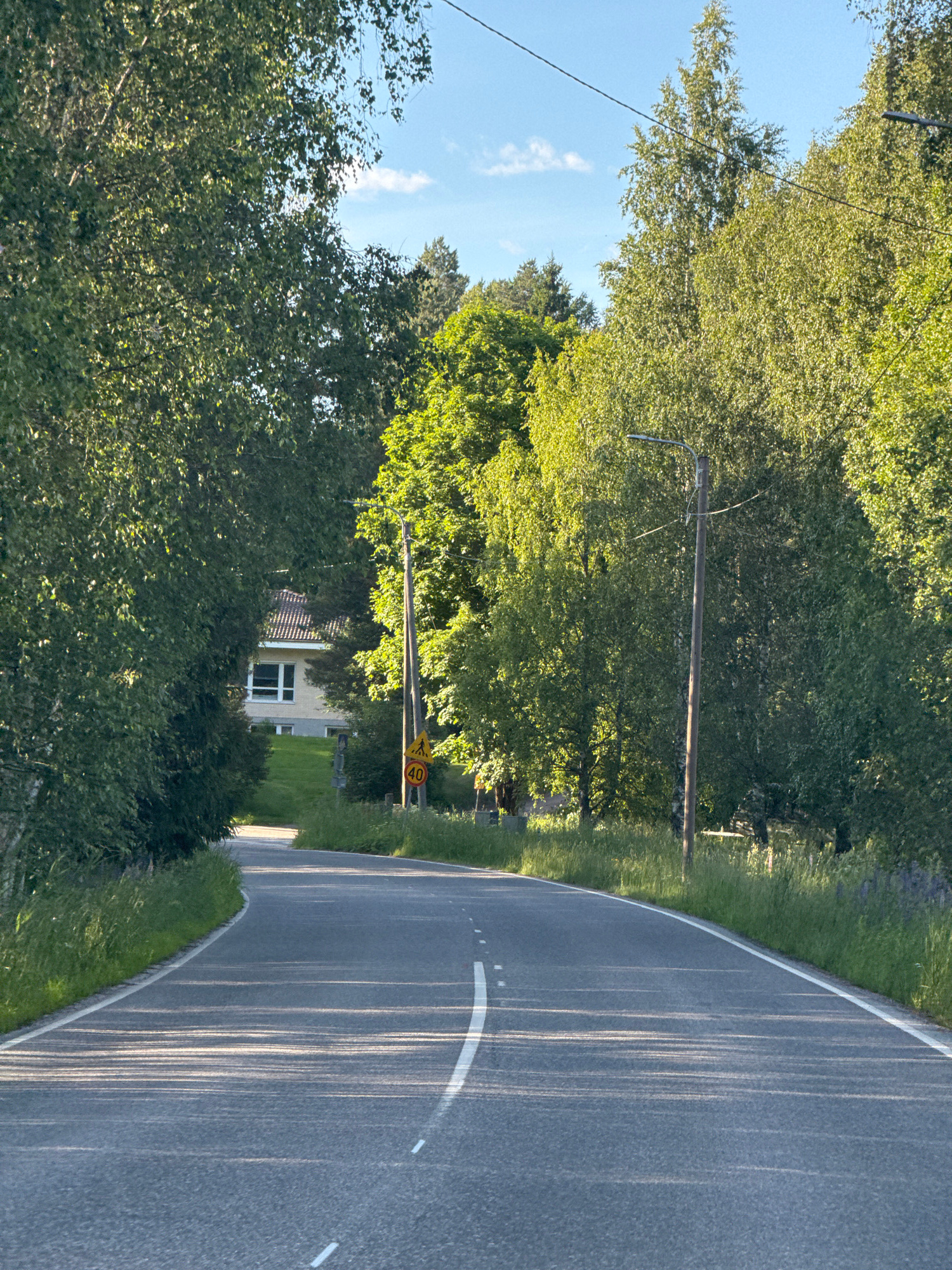 The image shows a paved road with a white dashed line and a solid white line marking the lanes. The road curves to the left and is bordered by grassy verges and trees on both sides. In the background, a yellow house with a brown tiled roof is visible, partially obscured by foliage. A wooden utility pole with a lamp and wires is present on the right side of the road. A yellow circular traffic sign with a pedestrian symbol and the number "40" is also visible on the right. The sky is blue with a few white clouds. The scene appears to be in a rural or suburban setting with ample greenery.