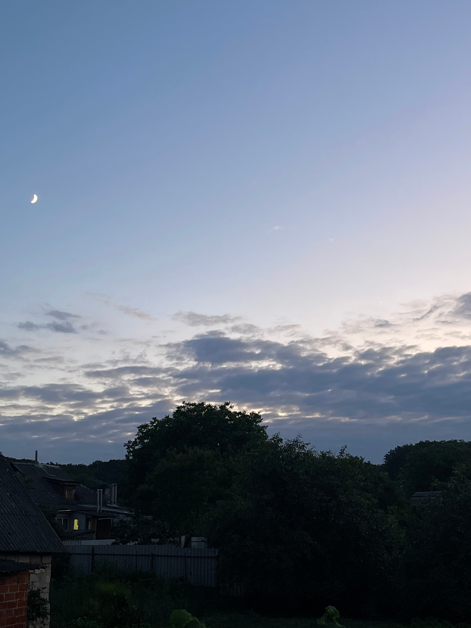 This is a vertical, medium shot taken from an elevated viewpoint, looking out over a rural or suburban landscape at dusk. The sky occupies the upper two-thirds of the frame and is a gradient of pale blue to light grey, with a sliver of the crescent moon visible in the upper left quadrant. A band of textured, grey clouds stretches across the middle ground of the sky.
The lower third of the image depicts a dense canopy of dark green trees, silhouetted against the brighter sky. In the left foreground, the dark, weathered roof of a building, possibly a house, is visible, constructed with slate tiles and some exposed brickwork. To the right of this building, another structure with a sloping, dark roof and visible chimneys is partially illuminated by a single, warm yellow light emanating from a window. A corrugated metal fence runs horizontally across the middle ground, separating the foreground from the background. The overall impression is one of a tranquil evening scene in the countryside near Krasnokutsk, Ukraine, with the transition from day to night.