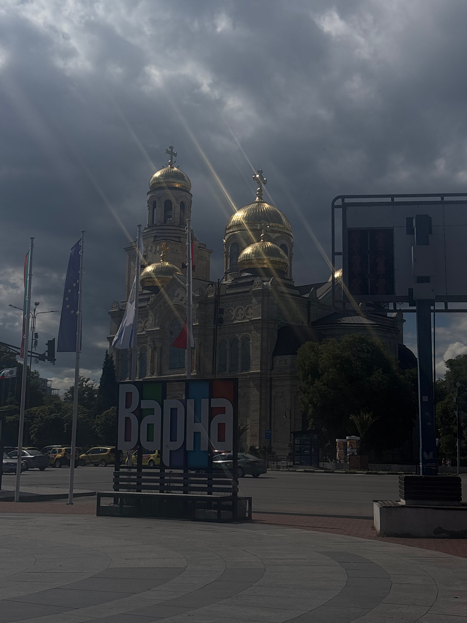 The image shows a cloudy sky with sun rays piercing through, over the Cathedral of the Assumption of the Virgin in Varna, Bulgaria. In the foreground, flags are displayed, including the Bulgarian flag, the European Union flag, and others. A large sign with colorful letters spells out "Варна" (Varna). Several cars are visible on the street, and trees line the background. The architecture of the cathedral, with its golden domes and crosses, is prominent. A bus stop and a street bench are also present.