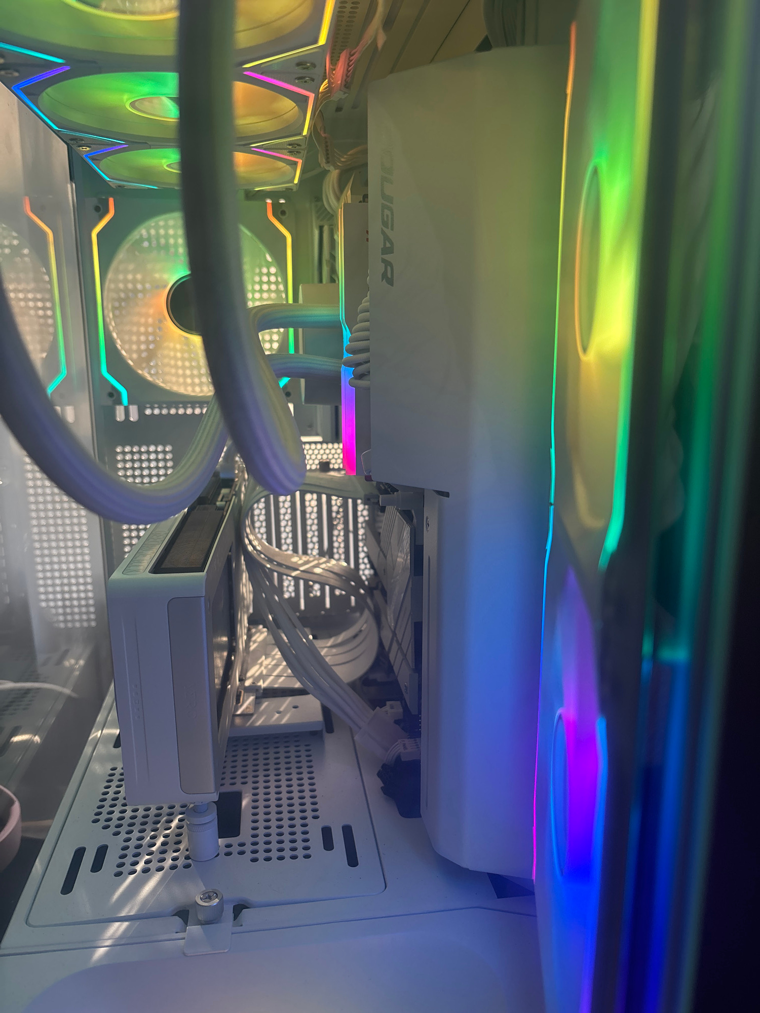 This is a close-up, low-angle shot inside a computer case. The primary objects are computer components, including fans, cables, and a white shroud with the "COUGAR" logo visible on it. The scene is illuminated by vibrant RGB lighting emanating from the fans and other internal components, casting a colorful glow across the white interior of the case. The lighting shifts through various colors, including green, yellow, blue, and purple.
The computer case appears to be white and features perforated panels, suggesting it is designed for airflow. Several white cables are neatly managed and routed throughout the case. A component, possibly a graphics card or an NVMe drive, is visible in the lower right portion of the image. The lighting creates reflections on the glass side panel of the case, adding depth to the image.
There is no discernible activity or event taking place, as the image focuses on the static components of the computer. The time of day and weather are not inferable from the image. The emotion conveyed is one of modernity and aesthetic appeal, often associated with enthusiast computer builds featuring RGB lighting. The visible text is "COUGAR".