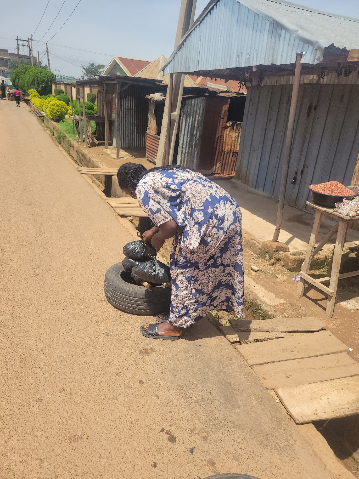 In Jos, Nigeria, a woman in a blue and white floral dress is seen standing on a paved road next to a ditch with wooden planks crossing it. She is bent over, tending to several black plastic bags of what appears to be trash placed inside an old tire. The scene is lined with small stalls made of corrugated metal and wood, with some displaying goods like grains and bagged items. Vegetation, including manicured green bushes and some trees, is visible along the roadside. The weather appears to be sunny and clear.